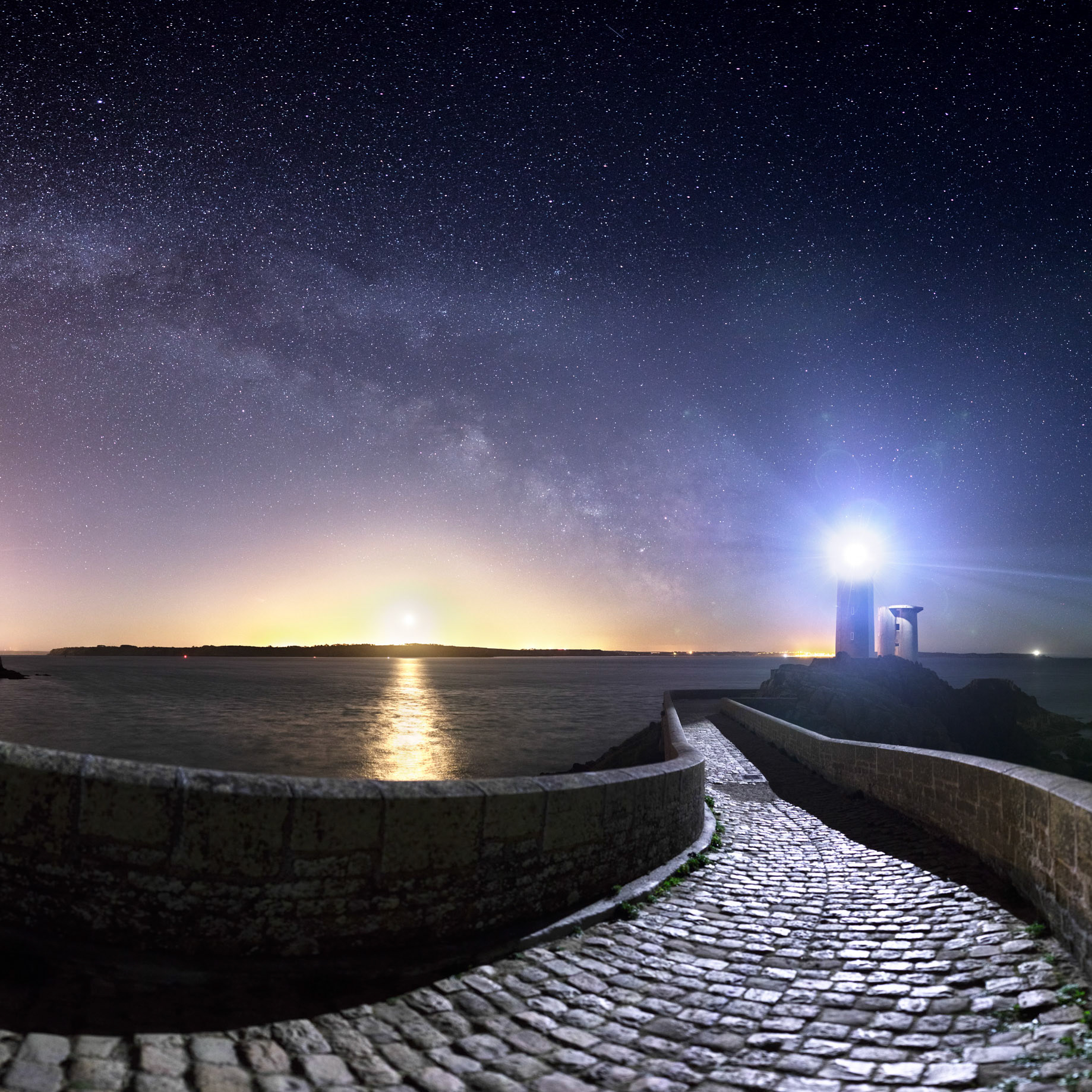 Le Quartier de Lune qui se lève sous la Voie lactée teinte le ciel en bleu, couleur qui s'oppose à la olution lumineuse qui entoure le phare du Petit Minou à Plouzané.