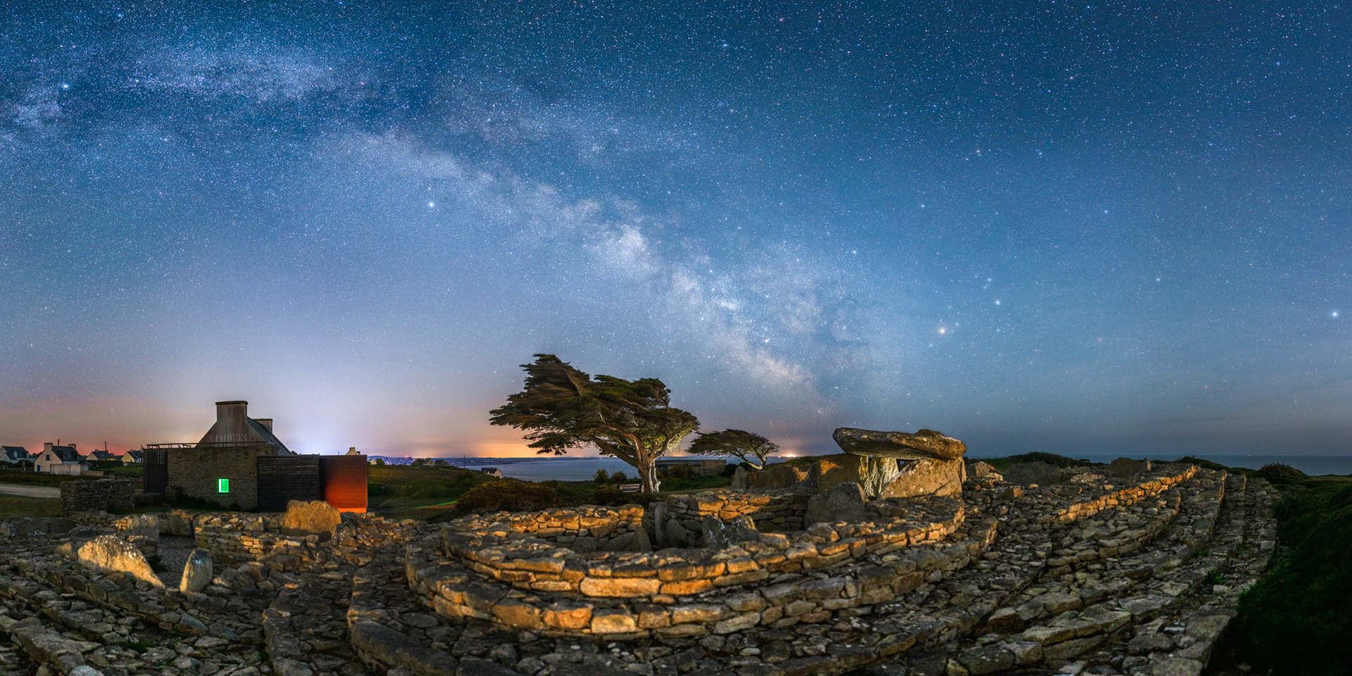Une allée couverte est érigée sur le site de Menez Dregan. De magnifiques arbres en drapeau semblent plier sous le poids de la Voie lactée qui barre le ciel. Pour que le relief soit bien visible, j'ai illuminé les rochers avec le flash de mon smartphone. Idem pour les arbres dont on perçoit chaque détail. Notez la lumière juste à droite de l'arbre de droite ; Il s'agit de Penmarc'h et de son phare. La lumière verte qui filtre à travers la fenêtre du bâtiment provient d'un bloc d'issue de secours !