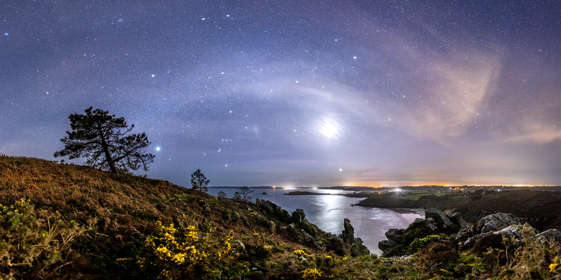 Le paysage qui s'offre à nous depuis la Pointe du Guern est des plus beaux. L'Île de l'Aber détache sa silhouette sombre sur le fond lumineux du reflet d'un Croissant de Lune. Juste au-dessus de l'îlot, le halo lumineux correspond au Port de Morgat (et son phare un peu plus haut à gauche). Dans le ciel, les nuages et le crépuscule masquent un peu la Voie lactée mais les nuées diffusent la lumière des étoiles les plus brillantes et rendent les constellations très visibles, avec notamment Orion entre la Lune et le pin de gauche auquel l'étoile Sirius est presque collée. Notez la petite tache irisée à gauche de la Lune. C'est un parasélène (la lumière est réfractée par les cristaux de glace des cirrus). Les fleurs jaunes des ajoncs apportent une touche de couleur complémentaire au bleu du ciel.