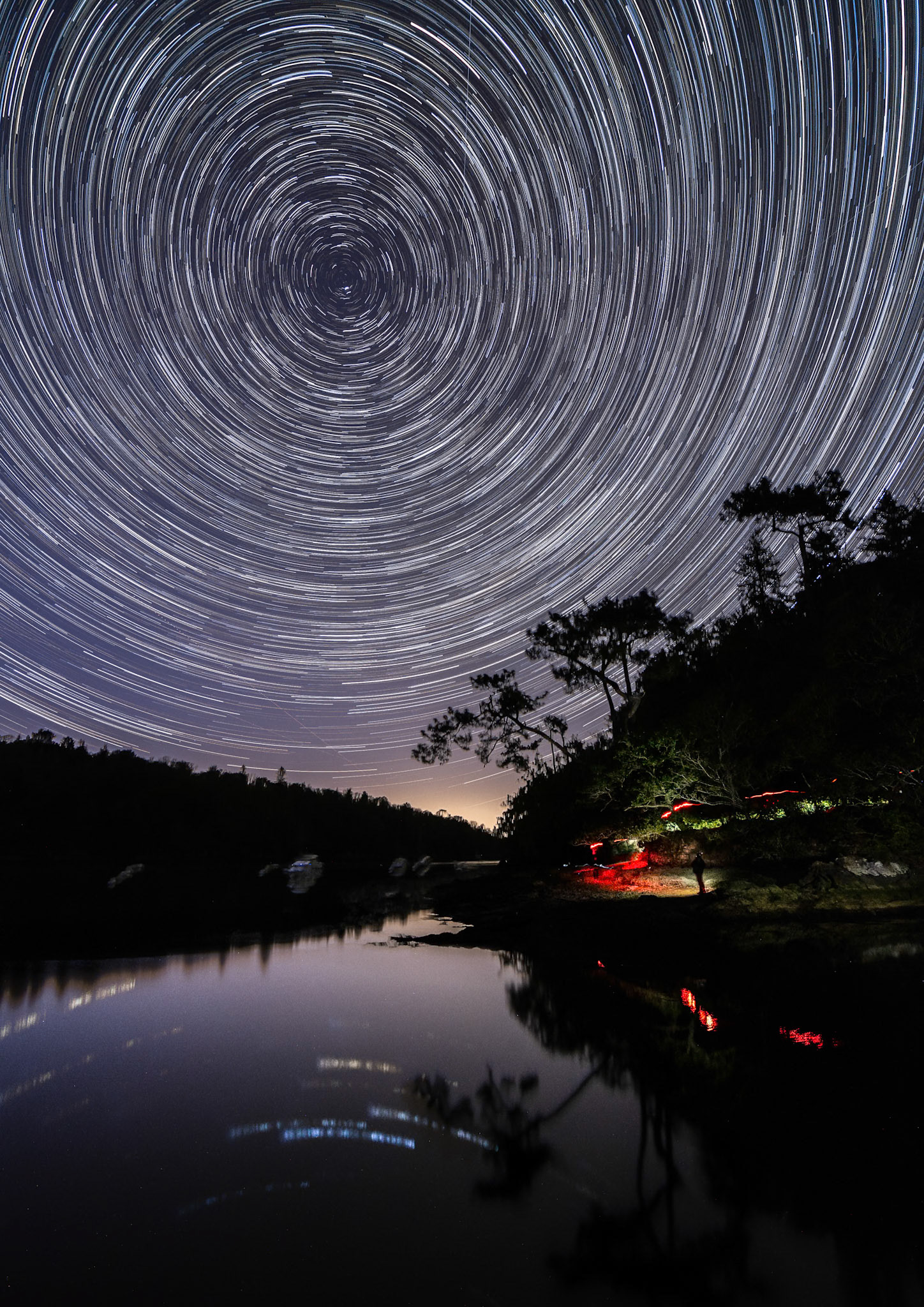 En réalisant 140 photos individuelles de 30 s chacune, je mets en évidence la rotation de la Terre de 23h40 à 0h55. Pour donner de la présence au coin inférieur droit, je suis allé éclairé la zone à l'aide du flash de mon smartphone et la lumière rouge de ma lampe frontale. Les grands pins qui se détachent au nord sur fond étoilé sont caractéristiques de Pors Meillou. Remarquez les étoiles les plus brillantes de la constellation de Cassiopée qui se reflettent dans les eaux calmes de l'Odet. La lueur orangée tout au fond est dû à la présence de Quimper.