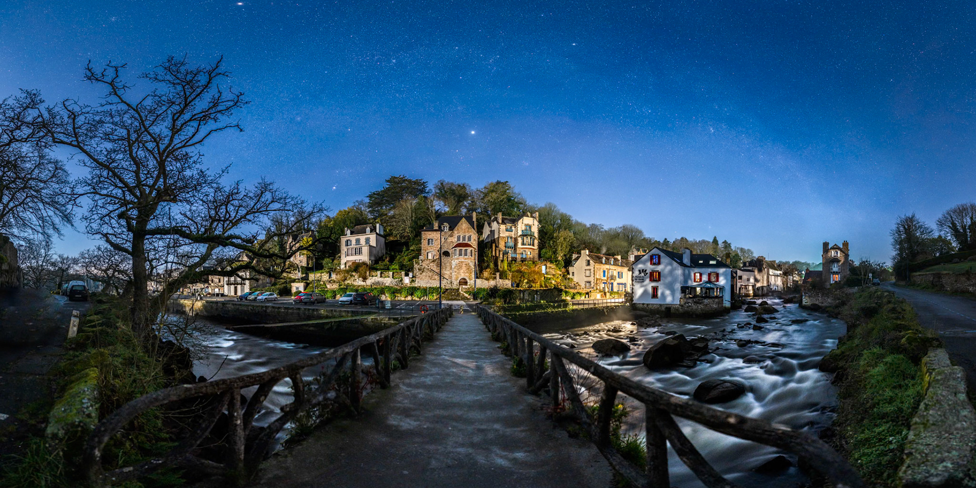 Le petit pont art déco qui enjambe la rivière Aven nous mène notre regard vers les jolies maisons de Pont-aven que surplombe la Voie lactée d'hiver. La Lune en Quartier illumine les collines sur la gauche tandis qu'une lumière orangée restée allumée sur le porche du restaurant le Moulin du Grand Poluguin teinte deux bâtiments d'une couleur chaude.