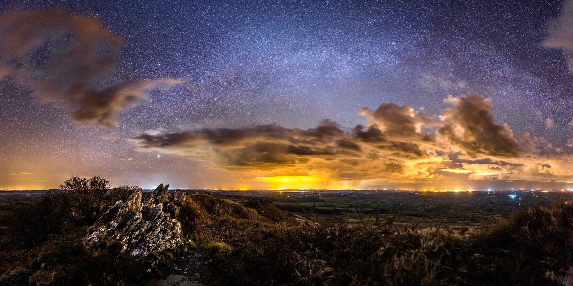 Les rochers en crête du Roc'h Trevezel se dressent à l'assaut du ciel où s'épanouit une belle Voie lactée malgré la pollution lumineuse forte (Brest est la source orange visible au milieu).