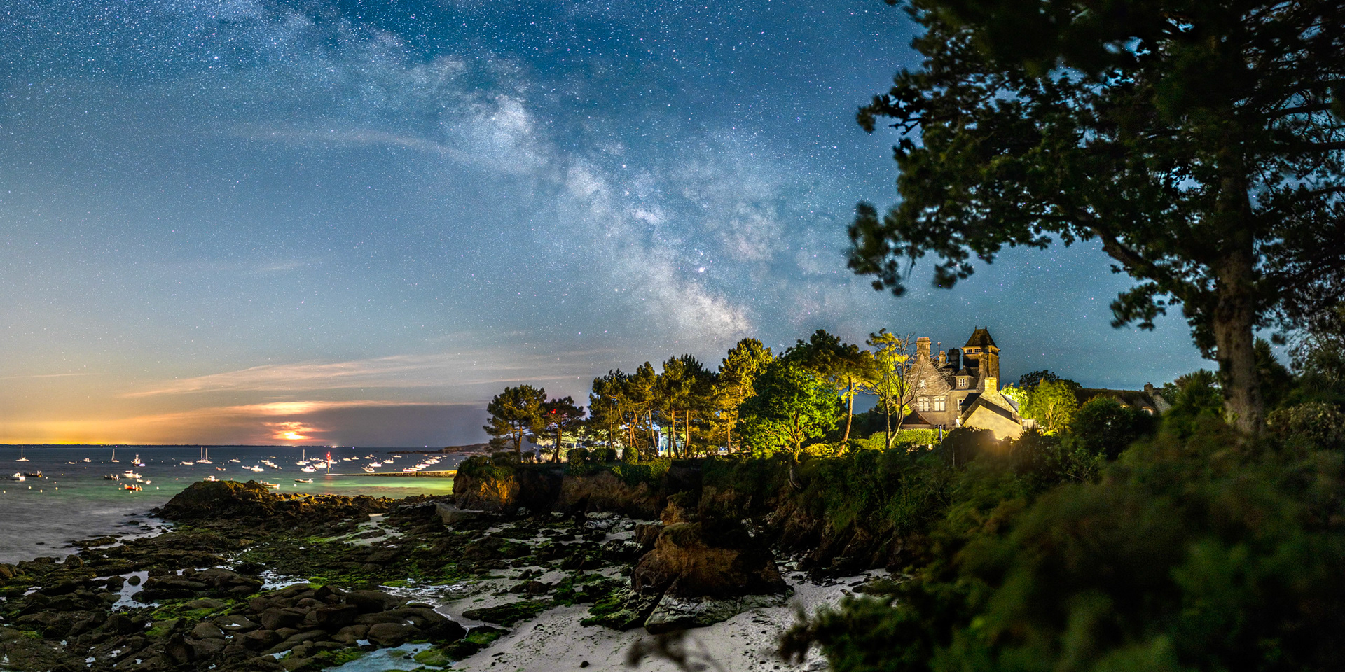 Le Manoir de Lanroz est magnifiquement illuminé, avec en fond la Voie lactée, mais aussi l'éclairage public qui illumine la cale de Beg Meil et donne sa teinte verte au fond sableux. A gauche, la présence orangée de Lorient et de la Lune (oblitérée par des nuages).