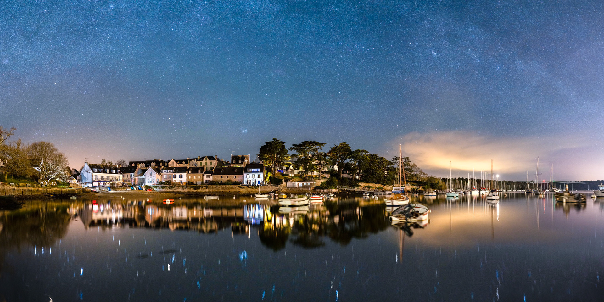 A marée haute, la mer n'est plus qu'un miroir où se reflètent les étoiles brillantes de la Voie lactée d'hiver qui forme une arche au-dessus du joli port de Sainte-Marine. A l'aide du flash de mon smartphone, j'ai éclairé les lieux les plus emblématiques du port :le chêne situé sous la chapelle, le café du port, l'abri du marin et le café de la cale. A droite, des cirrus qui planent au-dessus de Quimper apportent une jolie touche de lumière chaude.