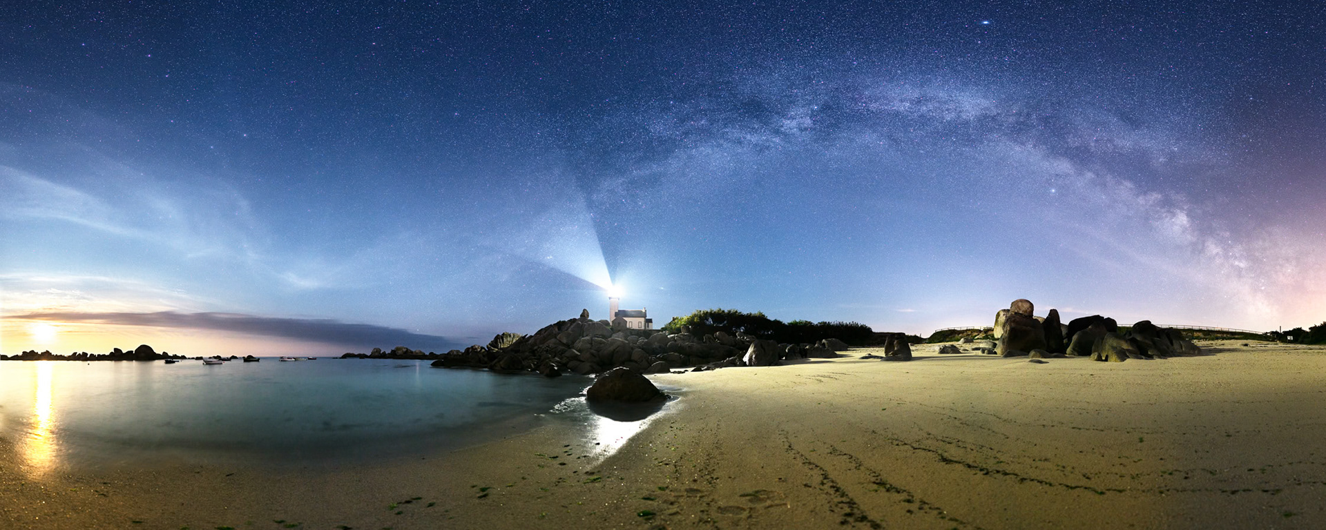 Tout à gauche, la Lune orange est en train de se coucher. Elle teinte encore le ciel de bleu où s'épanouit l'arche de la Voie lactée qui semble émerger du phare de Pontusval sur la plage de Brignogan.