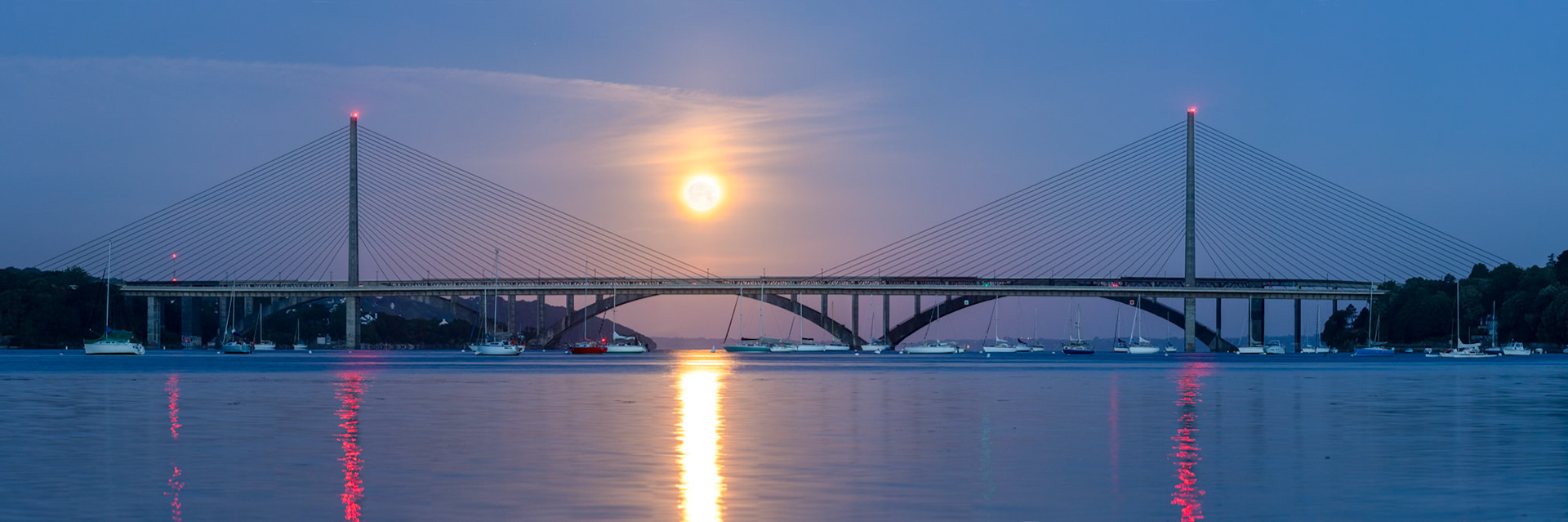 En juin, vu depuis les environs du centre nautique du Relecq-Kerhuon, la Pleine Lune se couche derrière le pont de l'Iroise. Les eaux de l'Elorn étant très calmes, les reflets des lumières au sommet des piles du pont sont magnifiques. Comme le jour est déjà bien levé, les haubans sombres sont parfiatement visibles sur fond clair.
