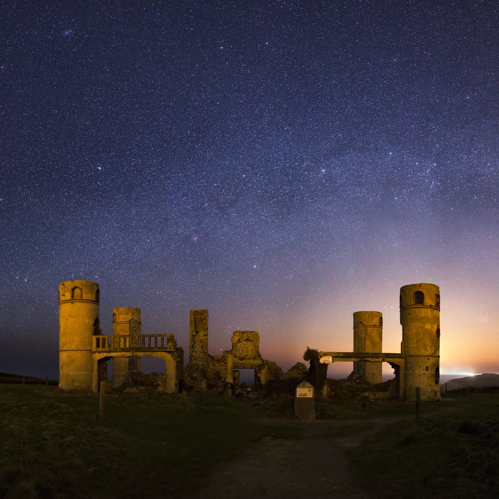 Les ruines du manoir de Saint-Pol-Roux sous l'arche de la Voie lactée, avec Orion qui se couche derrière le milieu des ruines. Le Quartier de Lune qui se couche derrière la tour de droite illumine l'horizon et bleuit le ciel.