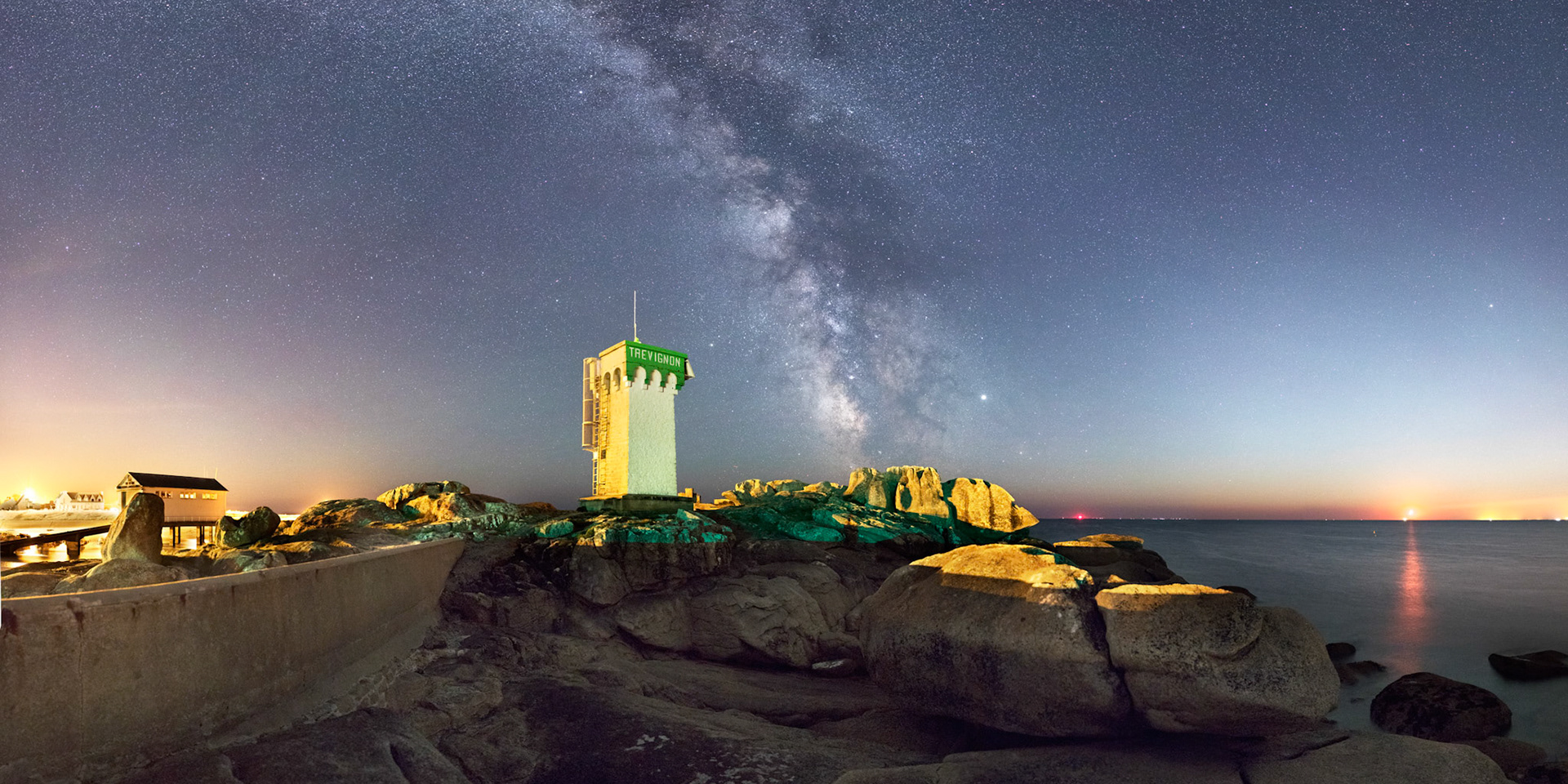 Tout à droite, la Lune se couche mais continue néanmoins à teinter le ciel en bleu. Différentes sources de lumière, lampadaires oranges, lampadaires blancs à LEDs et balise verte, contribuent à colorer le feu et les rochers de Trévignon où, dans le ciel, s'épanouit la Voie lactée.