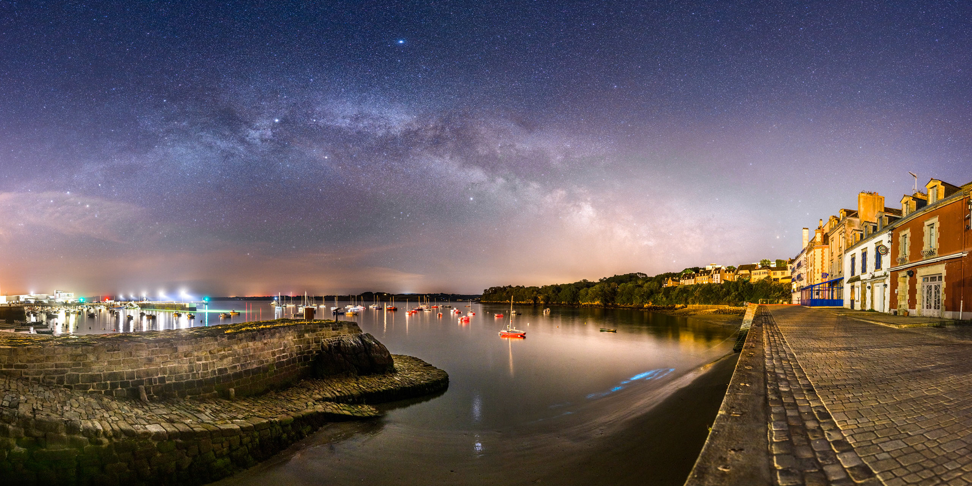 Depuis l'extinction des éclairages publics de Douarnenez, il est possible d'y faire de superbes photos. Le port de pêche situé tout à gauche offre avec ses lampadaires à LEDs une belle lumière neutre. Quelques balises rouges et vertes ajoutent des touches de couleur. Quant aux bateaux rouges situés au centre, la plupart d'entre eux sont blancs, mais illuminés par un feu rouge situé dans une rue qui donne sur le port (derrière la véranda bleue). Les traces de bleu en bas à droite sont dues au plancton phosphorescent. Et dans le ciel s'épanouit une superbe Voie lactée qui enjambe la cale ronde du port du Rosmeur et la baie de Douarnenez.