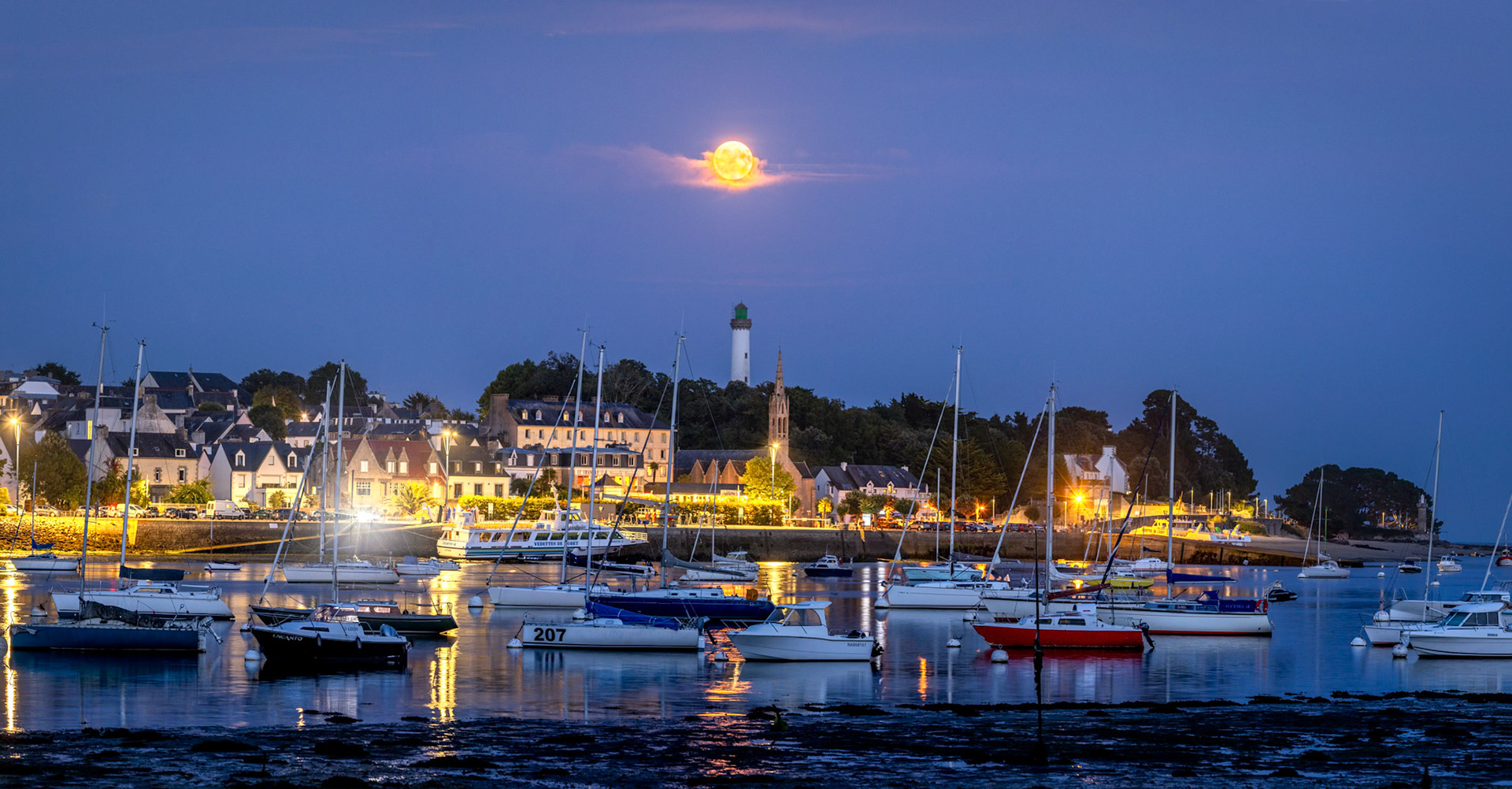La Pleine Lune semble posée sur un petit nuage à la verticale du phare de la Pyramide et de l'église. La lumère est superbe avec ce contraste entre les teintes orange de la Lune et des éclairages du port, et la teinte bleutée du crépuscule.