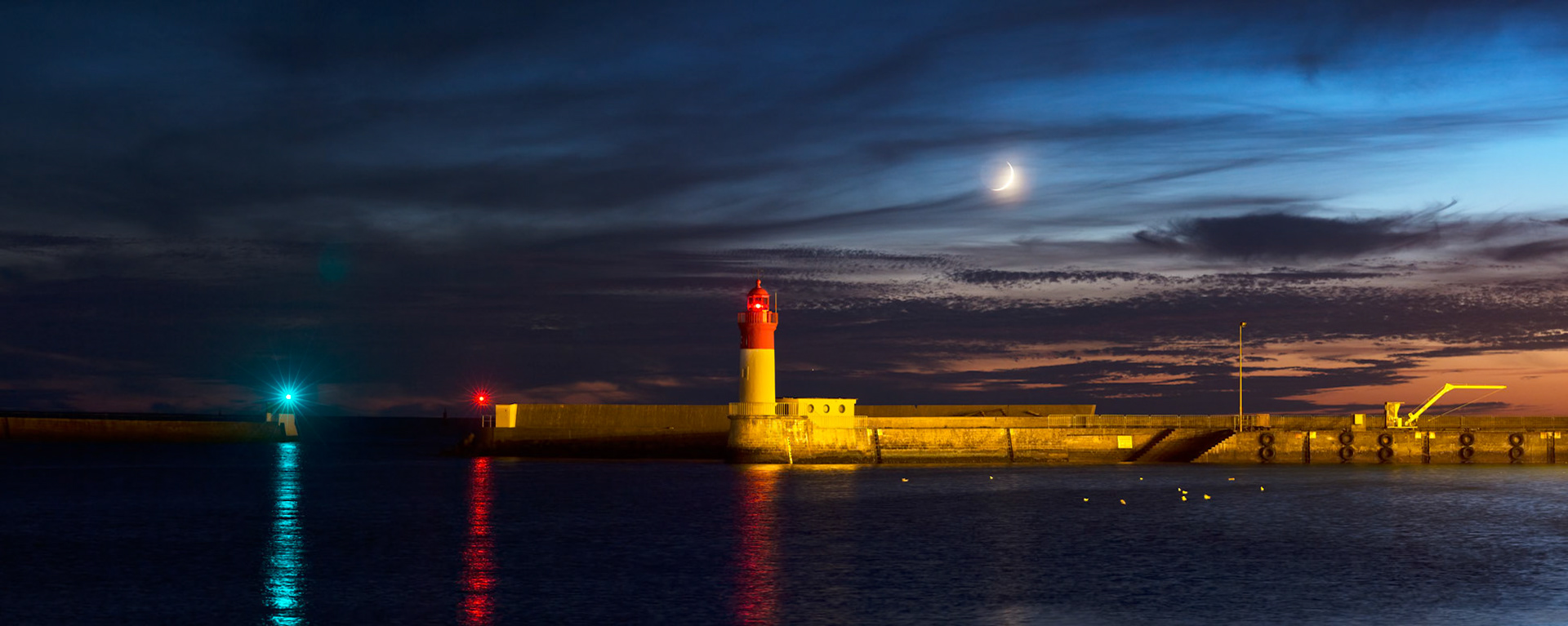 Le croissant de Lune se couche dans le crépuscule aux côtés du phare qui est dressé au bout d'une des digues du port. La discrète Vénus est située plus bas à gauche de la Lune.