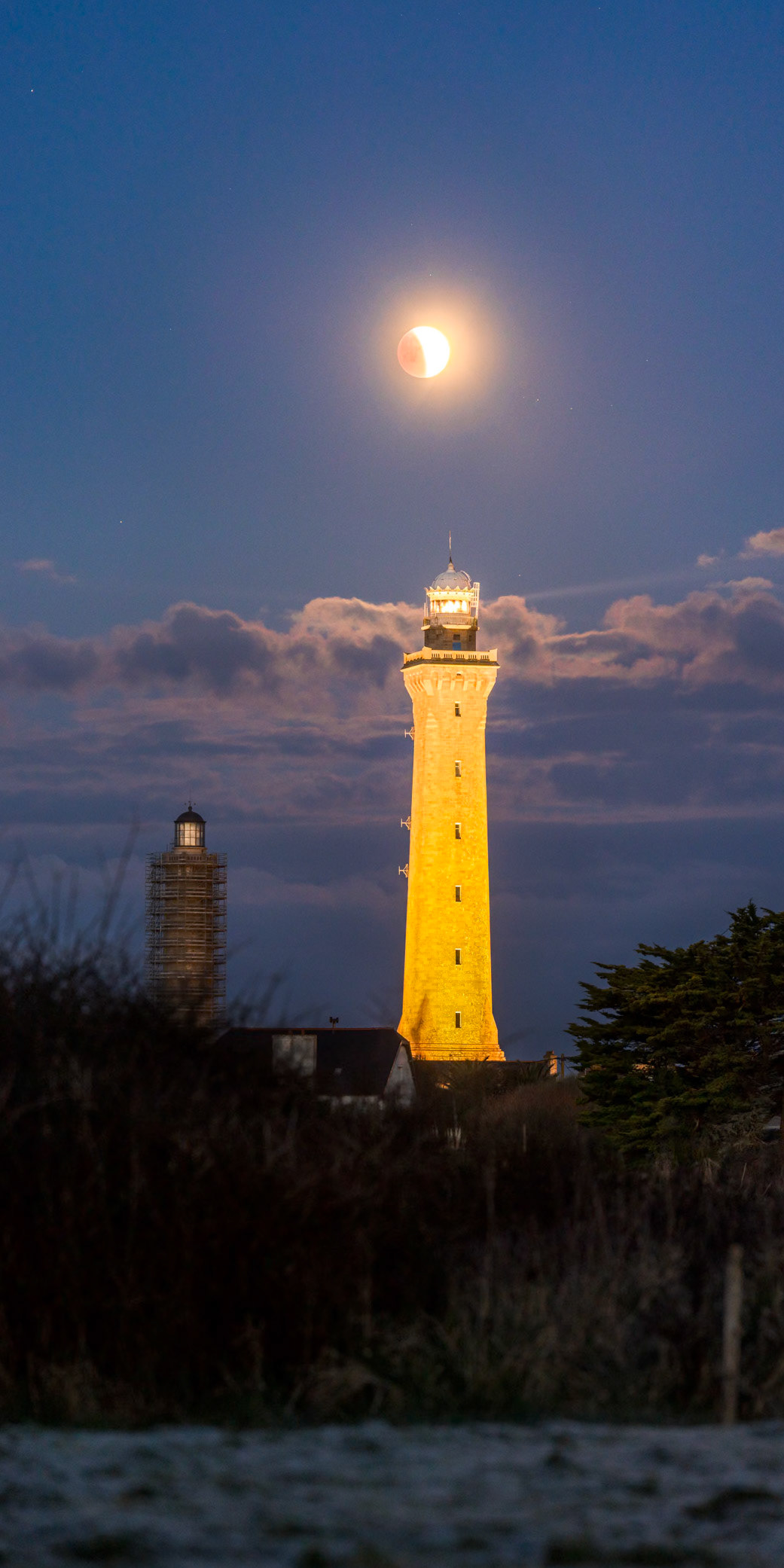 Installé dans un petit champ situé à l'est du phare d'Eckmühl, j'assiste à une éclipse de Lune qui n'est pour le moment que partielle. La phase totale ne sera pas visible car masquée par des nuages qui sont sur le point de cacher l'astre. Le corps du phare est éclairé de jaune-orangé qui contraste joliment avec le crépuscule bleuté. L'ancien phare vient d'être équipé d'une coupôle pour des raisons esthétiques mais son échaffaudage n'a pas encore été démonté.