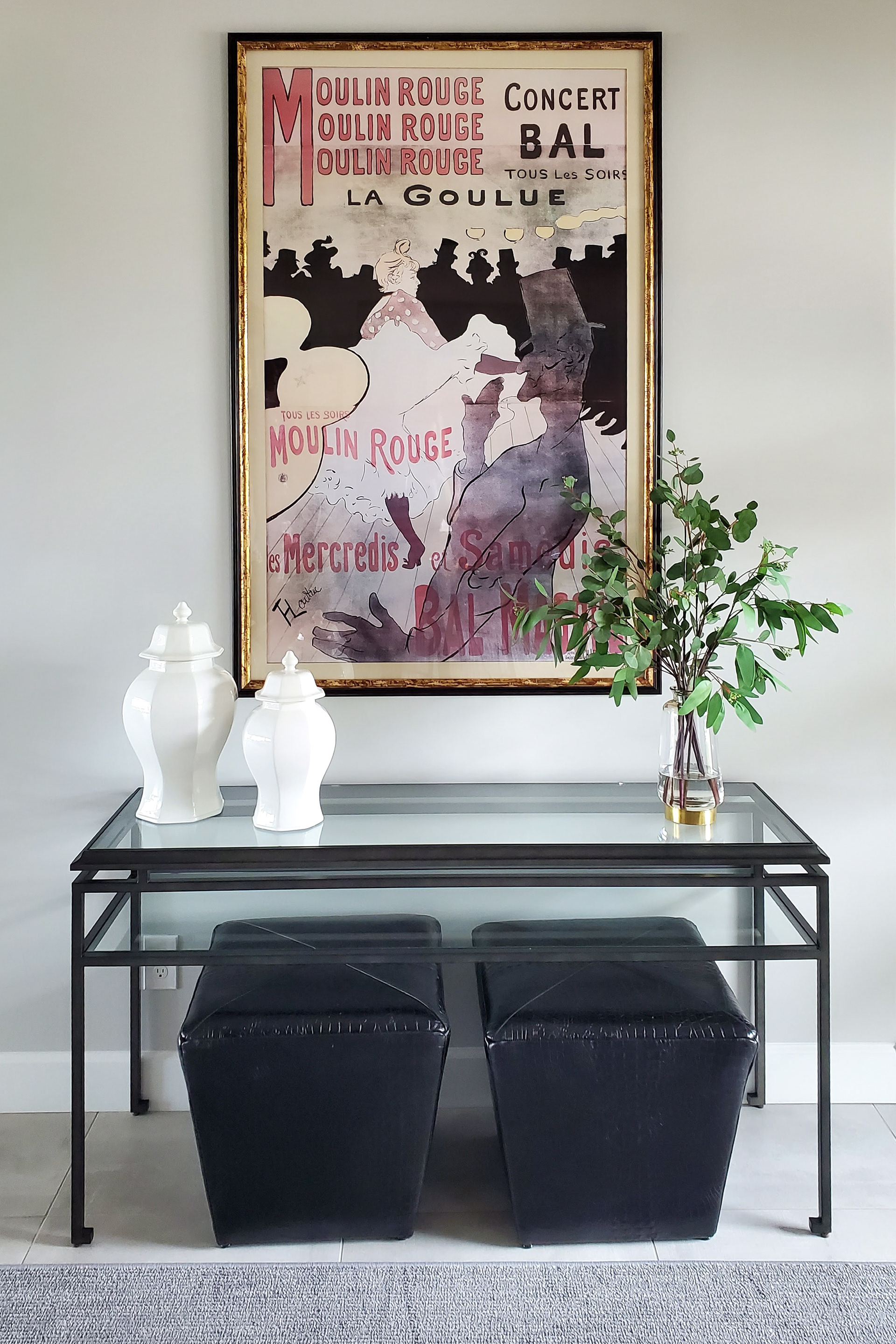 Interior design of a modern entry featuring a large vintage Moulin Rouge poster, a glass console table with black leather stools, and white ceramic ginger jars next to fresh greenery.