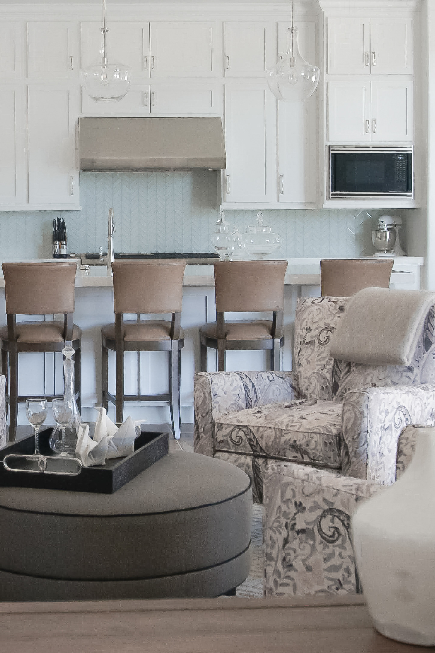 A bright, contemporary open-concept kitchen featuring white shaker cabinetry and light blue herringbone tile backsplash, transitioning into a neutral-toned lounge area with patterned armchairs and a grey circular ottoman.