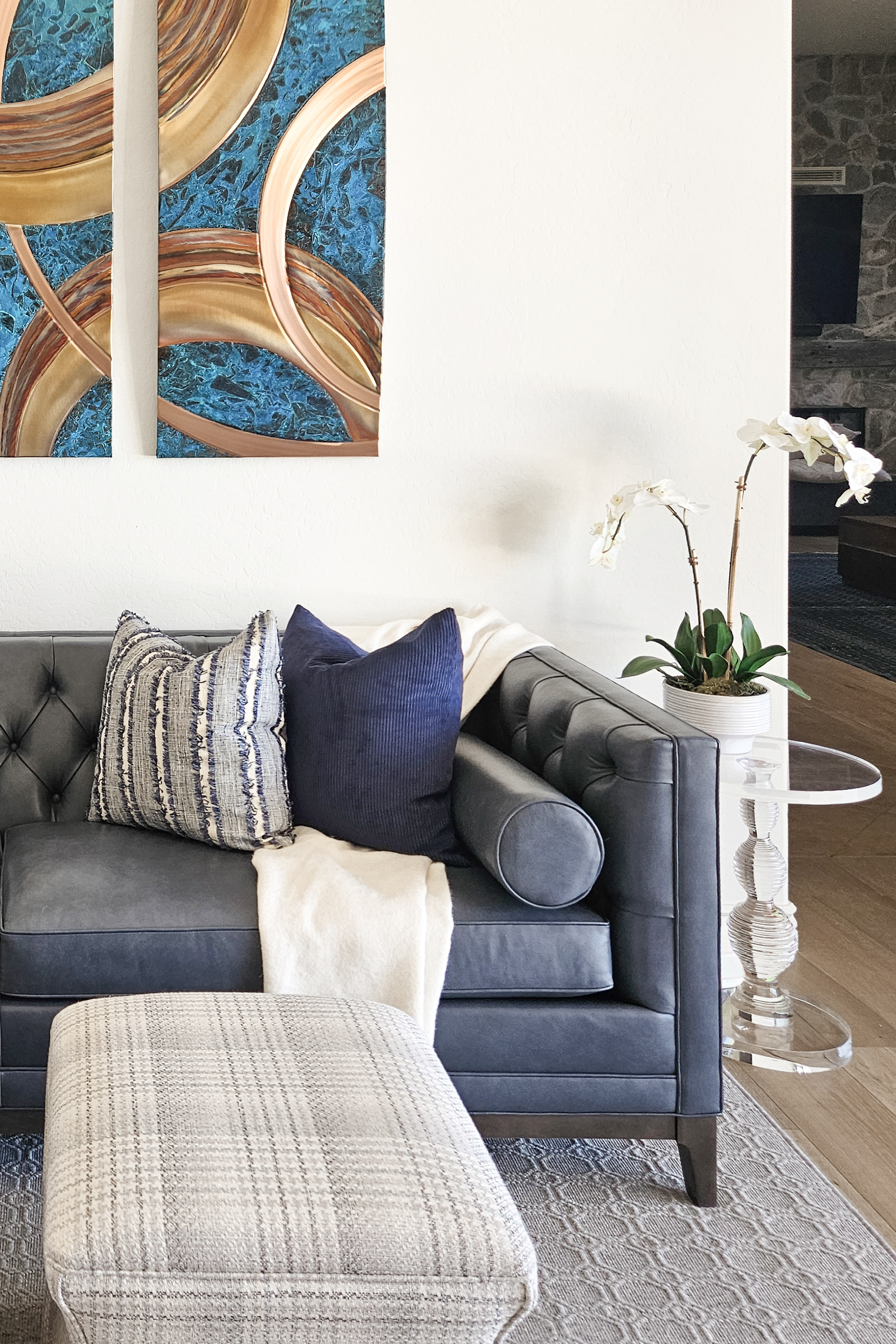Interior design of a luxury transitional living space featuring a deep slate blue tufted leather sofa, textured and corduroy throw pillows, and an ivory plaid ottoman. A modern glass side table with an orchid and abstract copper and blue triptych wall art complement the organic masonry fireplace in the background.