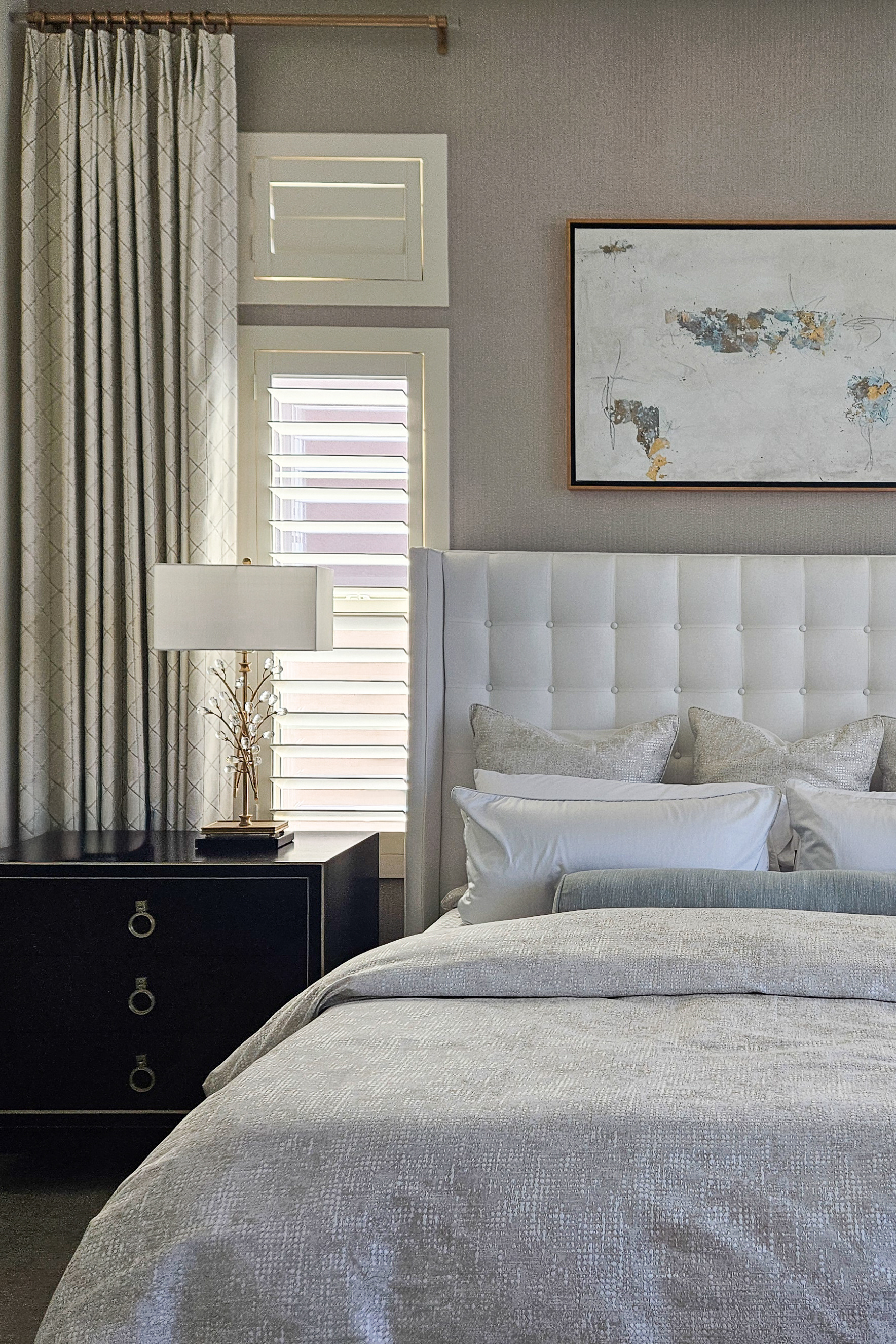 Interior design of a luxury primary bedroom featuring a cream tufted wingback headboard, shimmering champagne bedding, and an indonesian mahogany nightstand with gold ring pulls. A decorative gold branch lamp with crystal accents sits before a window with white shutters and geometric patterned drapes.