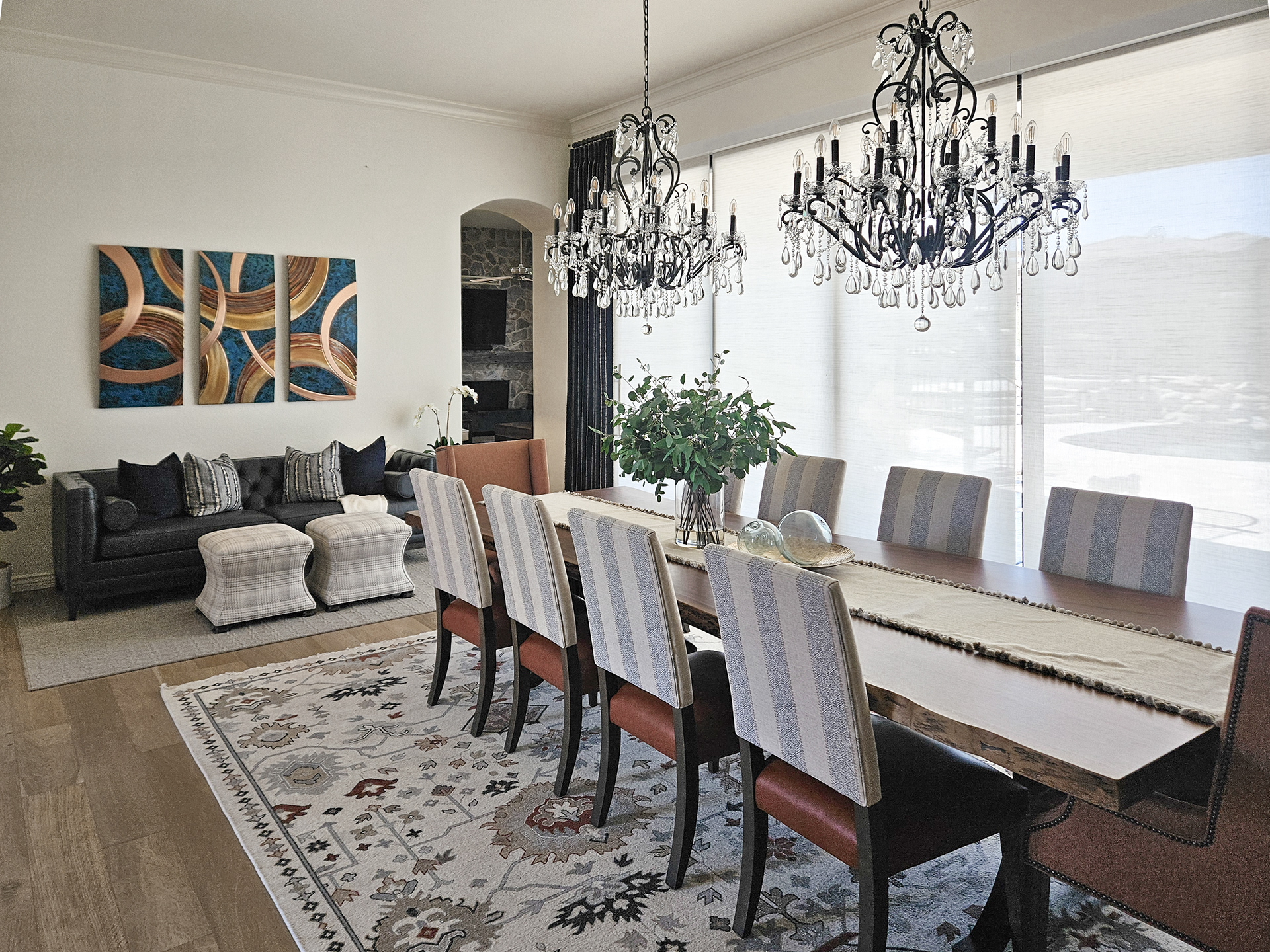 The Ember Nomad project featuring a long, dark wood live-edge dining table centered under ornate crystal chandeliers, surrounded by blue and white striped chairs and rust-colored end chairs within an open-concept flow.