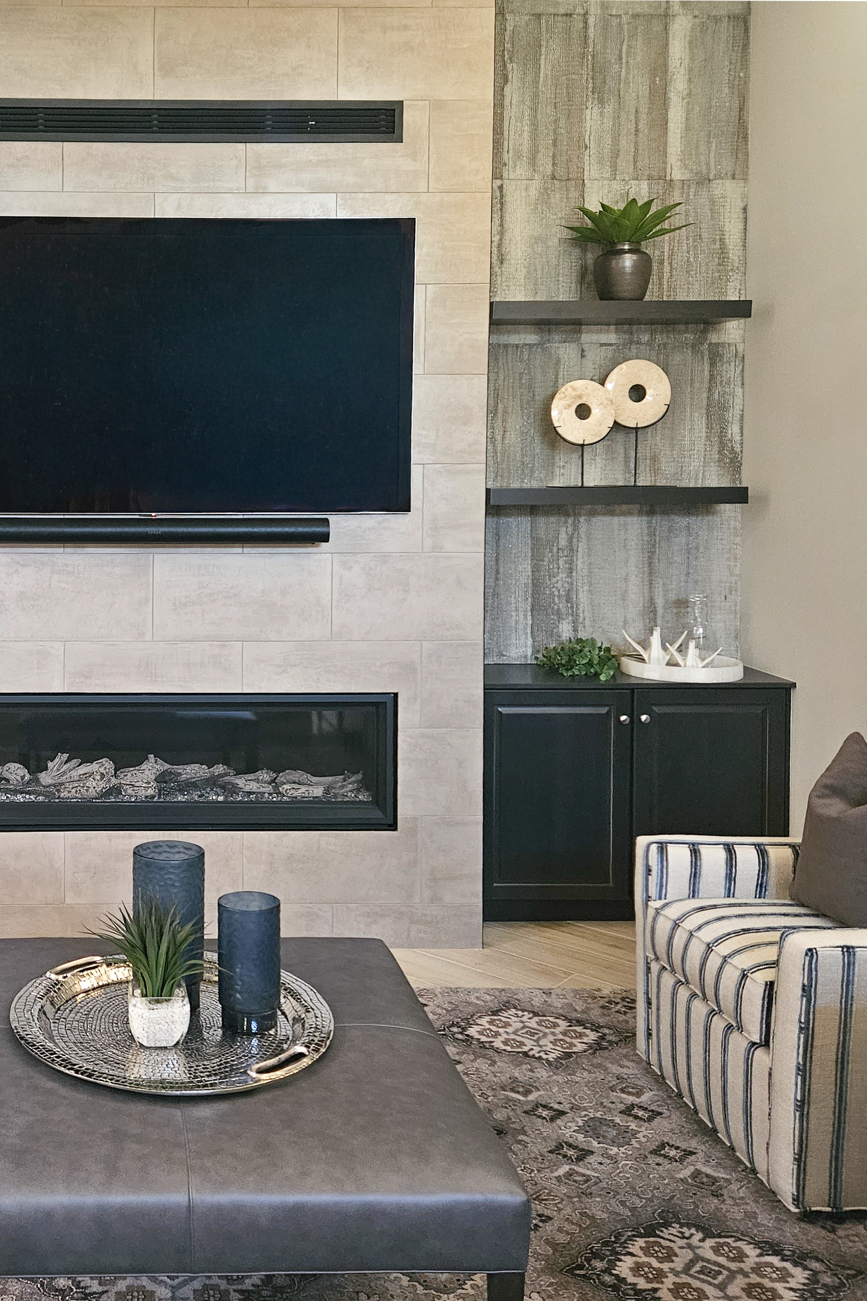 Interior design of a transitional living room featuring a large-format tiled fireplace, black built-in cabinetry, floating black shelves against a weathered wood-look accent wall, and a blue striped club chair.