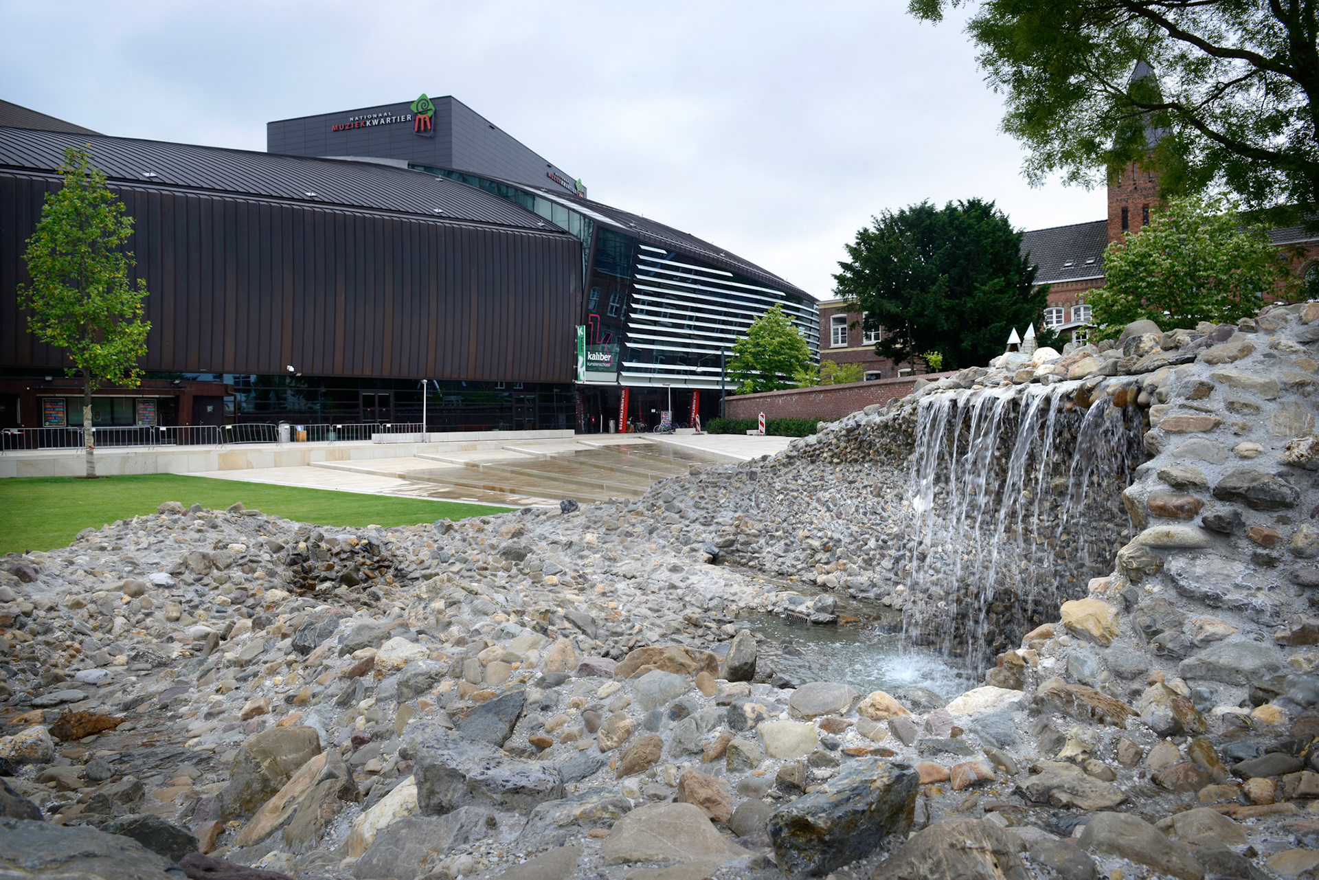 NIEUWE STADSTUIN VOOR ENSCHEDEWoensdag 18 juni zette wethouder Hatenboer de kraan open van het kunstwerk Vortex, onderdeel van het nieuwe Willem Wilminkplein in het centrum van Enschede. Sindsdien stroomt het water over een brede, luie natuurstenen trap richting de Vortex. Het kunstwerk is een uit keien opgebouwde ÔdraaikolkÕ waarin een waterval over een muurtje drie meter omlaag klatert.Kunstenaar Jeroen Doorenweerd ontwierp in nauwe samenwerking met gemeentelijk ontwerper Hans Schršder deze eigentijdse folly als integraal onderdeel van de trap en het plein. Hiermee heeft Enschede een sfeervolle groene huiskamer erbij gekregen.Komende zomermaanden kan het publiek het plein en het kunstwerk testen op hun recreatieve kwaliteiten. Op vrijdag 19 september vindt vervolgens de offici‘le opening van het plein plaats met een feestelijke act als startsein van het Willem Wilminkfestival.Muziekkwartier met Willem WilminkpleinIn het Muziekkwartier van Enschede zijn alle belangrijke muziekpodia- en gezelschappen van de stad te vinden. In het hart ervan ligt het nieuwe driehoekige Willem Wilminkplein. Historische gebouwen zoals een voormalig klooster en een fabrieksschool begrenzen samen met nieuwe gebouwen het plein en verlenen het karakter en intimiteit. Het Wilminktheater (2008) van Ector Hoogstad Architecten vormt met het nog op te leveren hotel van Dick van Gameren ŽŽn lange wand. Aan de overzijde realiseerde Onix architecten dit jaar een appartementencomplex met winkels en horeca.Het plein zal een oase in de stad zijn; slechts toegankelijk voor voetgangers en fietsers. Het grote grasveld, de banken eromheen en de cafŽterrassen nodigen uit om de ruimte als stadstuin te gebruiken. Aanlokkelijk middelpunt aan de oostkant vormt de waaiervormige luie trap waar water overheen stroomt, uitmondend in het kunstwerk Vortex. De trap kan gebruikt worden als podium voor kleinschalige muziekoptredens. Een andere blikvanger aan de westkant van het plein wordt de nog in aanbouw zijn