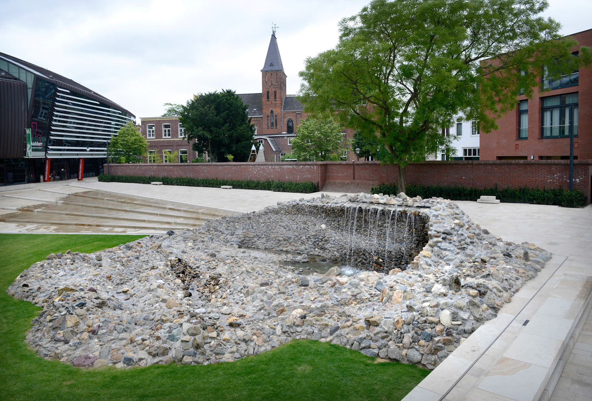 NIEUWE STADSTUIN VOOR ENSCHEDEWoensdag 18 juni zette wethouder Hatenboer de kraan open van het kunstwerk Vortex, onderdeel van het nieuwe Willem Wilminkplein in het centrum van Enschede. Sindsdien stroomt het water over een brede, luie natuurstenen trap richting de Vortex. Het kunstwerk is een uit keien opgebouwde ÔdraaikolkÕ waarin een waterval over een muurtje drie meter omlaag klatert.Kunstenaar Jeroen Doorenweerd ontwierp in nauwe samenwerking met gemeentelijk ontwerper Hans Schršder deze eigentijdse folly als integraal onderdeel van de trap en het plein. Hiermee heeft Enschede een sfeervolle groene huiskamer erbij gekregen.Komende zomermaanden kan het publiek het plein en het kunstwerk testen op hun recreatieve kwaliteiten. Op vrijdag 19 september vindt vervolgens de offici‘le opening van het plein plaats met een feestelijke act als startsein van het Willem Wilminkfestival.Muziekkwartier met Willem WilminkpleinIn het Muziekkwartier van Enschede zijn alle belangrijke muziekpodia- en gezelschappen van de stad te vinden. In het hart ervan ligt het nieuwe driehoekige Willem Wilminkplein. Historische gebouwen zoals een voormalig klooster en een fabrieksschool begrenzen samen met nieuwe gebouwen het plein en verlenen het karakter en intimiteit. Het Wilminktheater (2008) van Ector Hoogstad Architecten vormt met het nog op te leveren hotel van Dick van Gameren ŽŽn lange wand. Aan de overzijde realiseerde Onix architecten dit jaar een appartementencomplex met winkels en horeca.Het plein zal een oase in de stad zijn; slechts toegankelijk voor voetgangers en fietsers. Het grote grasveld, de banken eromheen en de cafŽterrassen nodigen uit om de ruimte als stadstuin te gebruiken. Aanlokkelijk middelpunt aan de oostkant vormt de waaiervormige luie trap waar water overheen stroomt, uitmondend in het kunstwerk Vortex. De trap kan gebruikt worden als podium voor kleinschalige muziekoptredens. Een andere blikvanger aan de westkant van het plein wordt de nog in aanbouw zijn