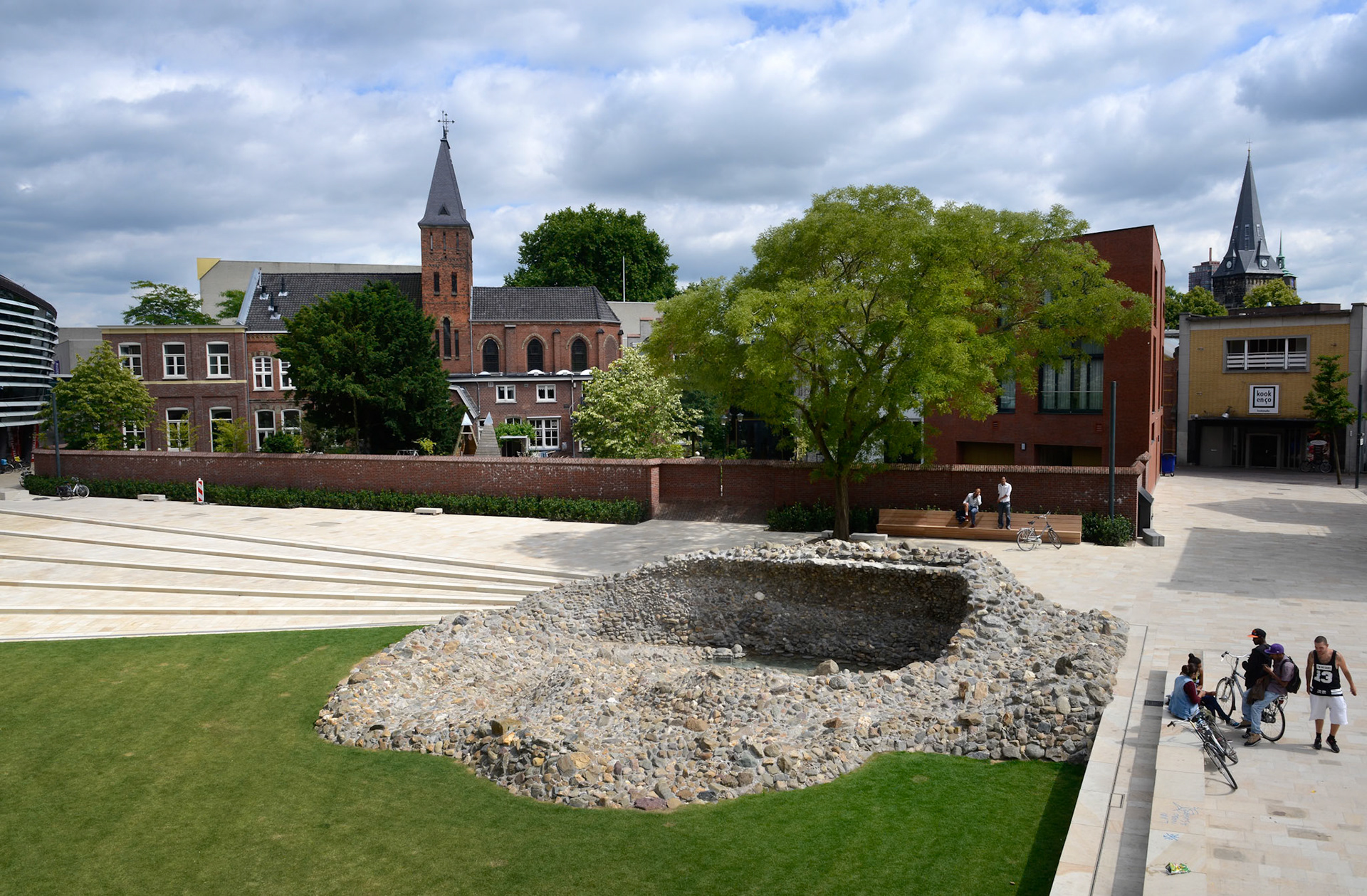 NIEUWE STADSTUIN VOOR ENSCHEDEWoensdag 18 juni zette wethouder Hatenboer de kraan open van het kunstwerk Vortex, onderdeel van het nieuwe Willem Wilminkplein in het centrum van Enschede. Sindsdien stroomt het water over een brede, luie natuurstenen trap richting de Vortex. Het kunstwerk is een uit keien opgebouwde ÔdraaikolkÕ waarin een waterval over een muurtje drie meter omlaag klatert.Kunstenaar Jeroen Doorenweerd ontwierp in nauwe samenwerking met gemeentelijk ontwerper Hans Schršder deze eigentijdse folly als integraal onderdeel van de trap en het plein. Hiermee heeft Enschede een sfeervolle groene huiskamer erbij gekregen.Komende zomermaanden kan het publiek het plein en het kunstwerk testen op hun recreatieve kwaliteiten. Op vrijdag 19 september vindt vervolgens de offici‘le opening van het plein plaats met een feestelijke act als startsein van het Willem Wilminkfestival.Muziekkwartier met Willem WilminkpleinIn het Muziekkwartier van Enschede zijn alle belangrijke muziekpodia- en gezelschappen van de stad te vinden. In het hart ervan ligt het nieuwe driehoekige Willem Wilminkplein. Historische gebouwen zoals een voormalig klooster en een fabrieksschool begrenzen samen met nieuwe gebouwen het plein en verlenen het karakter en intimiteit. Het Wilminktheater (2008) van Ector Hoogstad Architecten vormt met het nog op te leveren hotel van Dick van Gameren ŽŽn lange wand. Aan de overzijde realiseerde Onix architecten dit jaar een appartementencomplex met winkels en horeca.Het plein zal een oase in de stad zijn; slechts toegankelijk voor voetgangers en fietsers. Het grote grasveld, de banken eromheen en de cafŽterrassen nodigen uit om de ruimte als stadstuin te gebruiken. Aanlokkelijk middelpunt aan de oostkant vormt de waaiervormige luie trap waar water overheen stroomt, uitmondend in het kunstwerk Vortex. De trap kan gebruikt worden als podium voor kleinschalige muziekoptredens. Een andere blikvanger aan de westkant van het plein wordt de nog in aanbouw zijnde monumentale buitentrap die toegang zal geven tot de muziekschool in het Wilminktheater en het nieuwe hotel. Ook deze trap kan als podium en verblijfsplek dienen en biedt bovendien uitzicht op het naastgelegen stationsplein.VortexDe vorm van het plein inspireerde kunstenaar Jeroen Doorenweerd (Tilburg) tot zijn kunstwerk. Hij vertelt: ÒHet plein is driehoekig en door de diagonalen krijgt de ruimte dynamiek. De treden van de trap zijn samengeknepen lijnen die op het smalste punt een natuurlijke aanleiding vormen voor een cirkel, waarin de Vortex ligt. Over een wandje klatert water drie meter omlaag als een gecultiveerde waterval. Via stapstenen kun je, als je durft, naar beneden lopen, achter het water langs. De vormgeving met keien is afgeleid van de techniek van de gewassen grindtegel, die in de jaren zeventig grote populariteit genoot.ÓDoorenweerd over de inhoud van zijn werk: ÓVortex betekent eigenlijk werveling, in vloeistof of in lucht, zoals een draaikolk of een tornado. Daarnaast is het ook een term die veel in de Science Fiction wordt gebruikt; Ôto disappear into the vortexÕ wil zeggen dat je in een andere tijd en ruimte terecht komt. Je wordt tegen wil en dank meegenomen naar een andere wereld en daar ligt een parallel met kunst, want kunst behoort dat eigenlijk ook te doen.Ó©foto eric brinkhorst