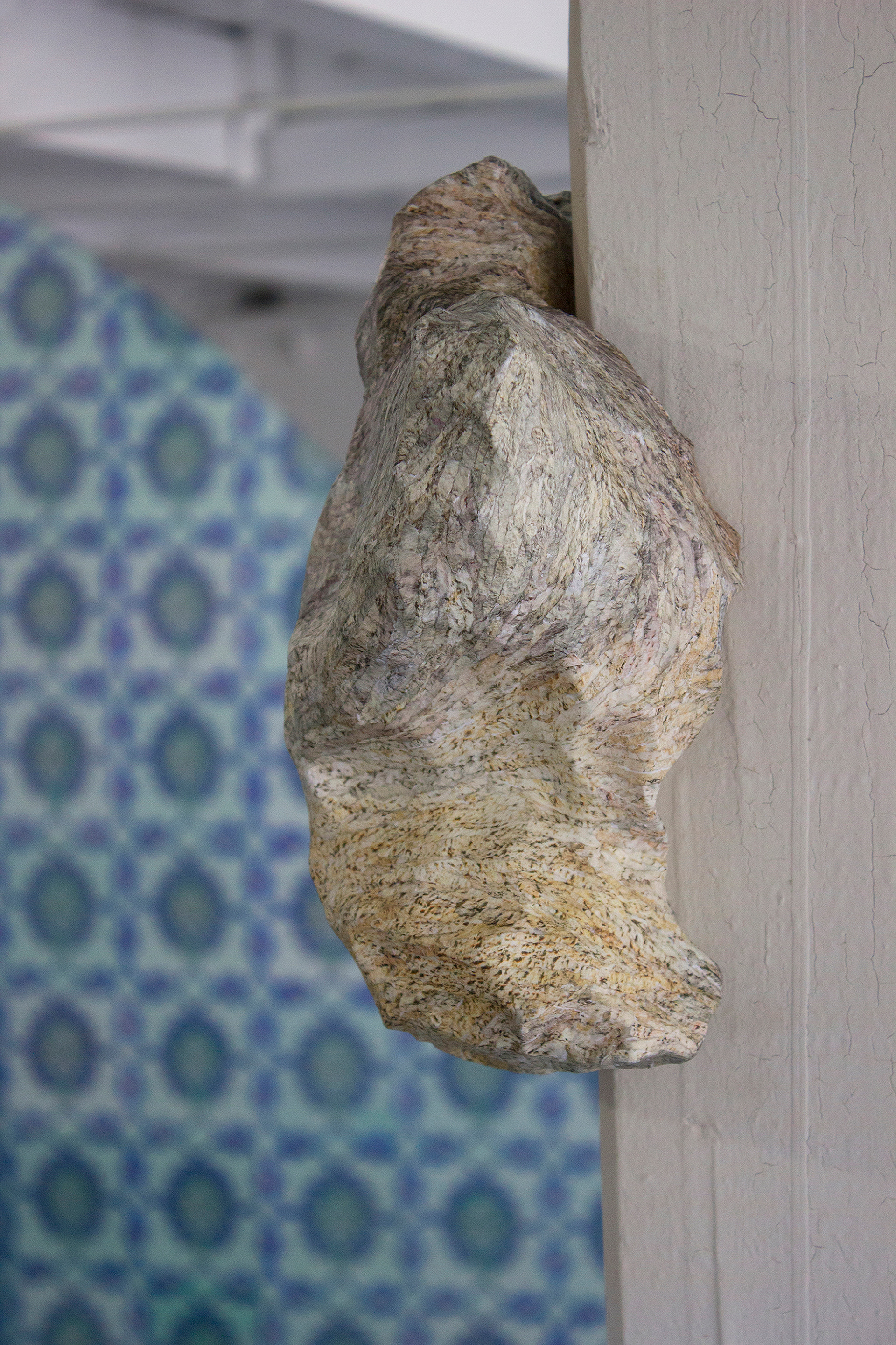 "Their Natural State" Rubble sculpture detail showing various shades of tan and grey blending together to create a rock texture. The sculpture hangs on one of the gallery's columns, with a tile archway appearing in the distance