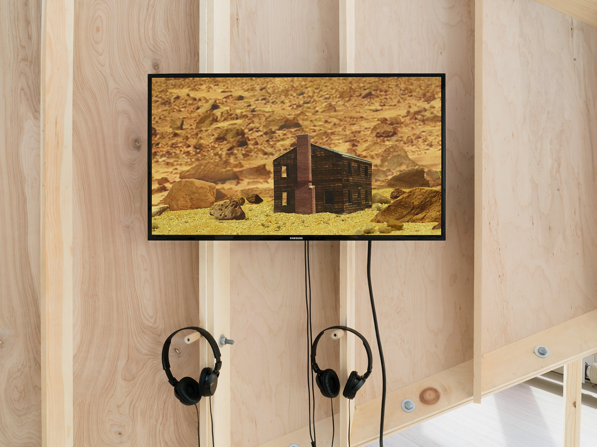 Image description: A TV monitor hangs on the outside of the sculpture "Silver Bullet Test Site" with headphones also dangling from the sculpture. The monitor shows the image of a lone abandoned wooden house in the middle of the desert. The image is created using found online images that are cut and assembled for the video.