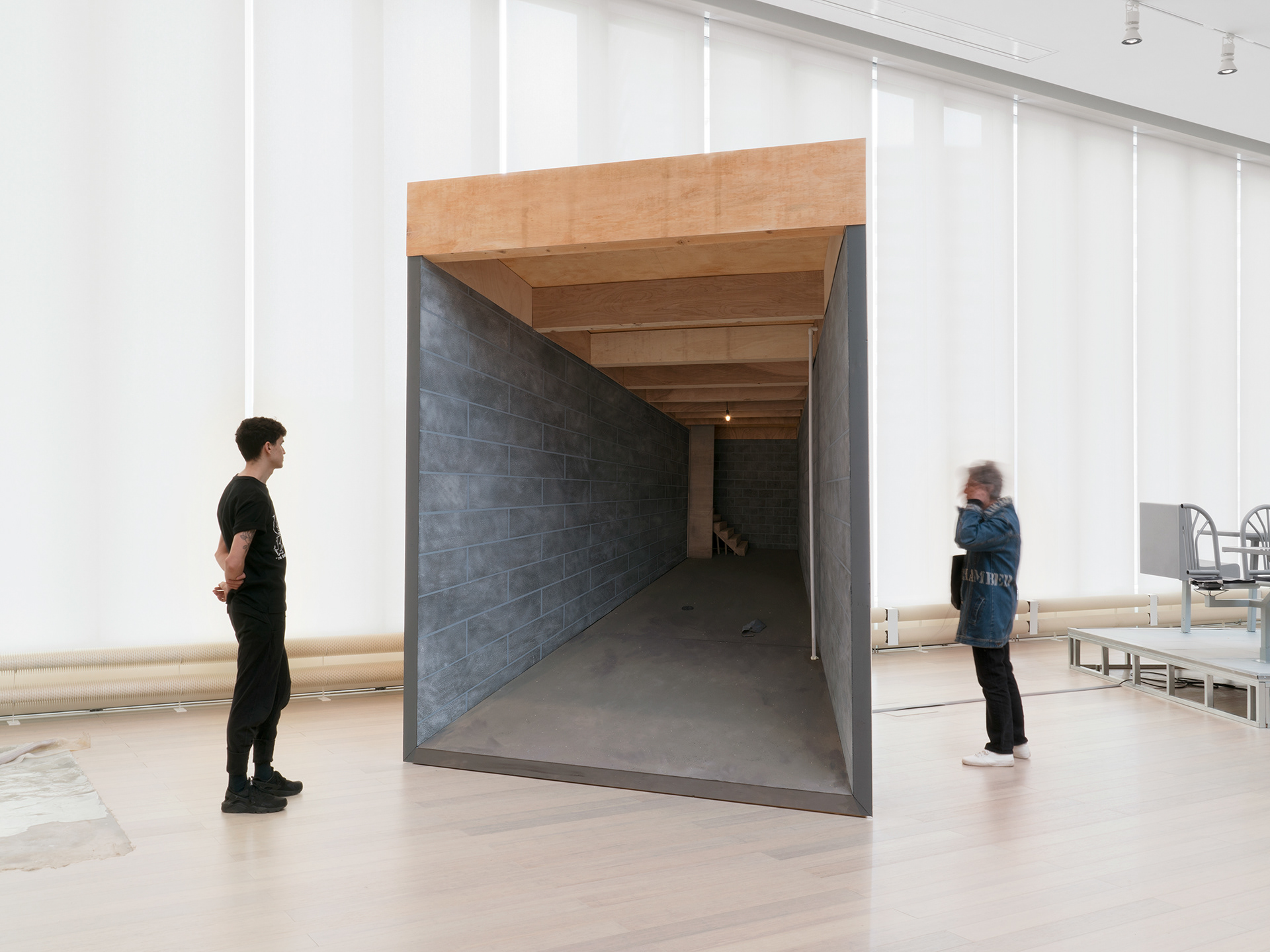 "Silver Bullet Test Site" overall shot (shades closed) with two viewers interacting with the sculpture: one stands in front contemplating the entry to a fictional basement room, and the other to the side of the sculpture, using headphones and watching the video on the exterior of the structure.