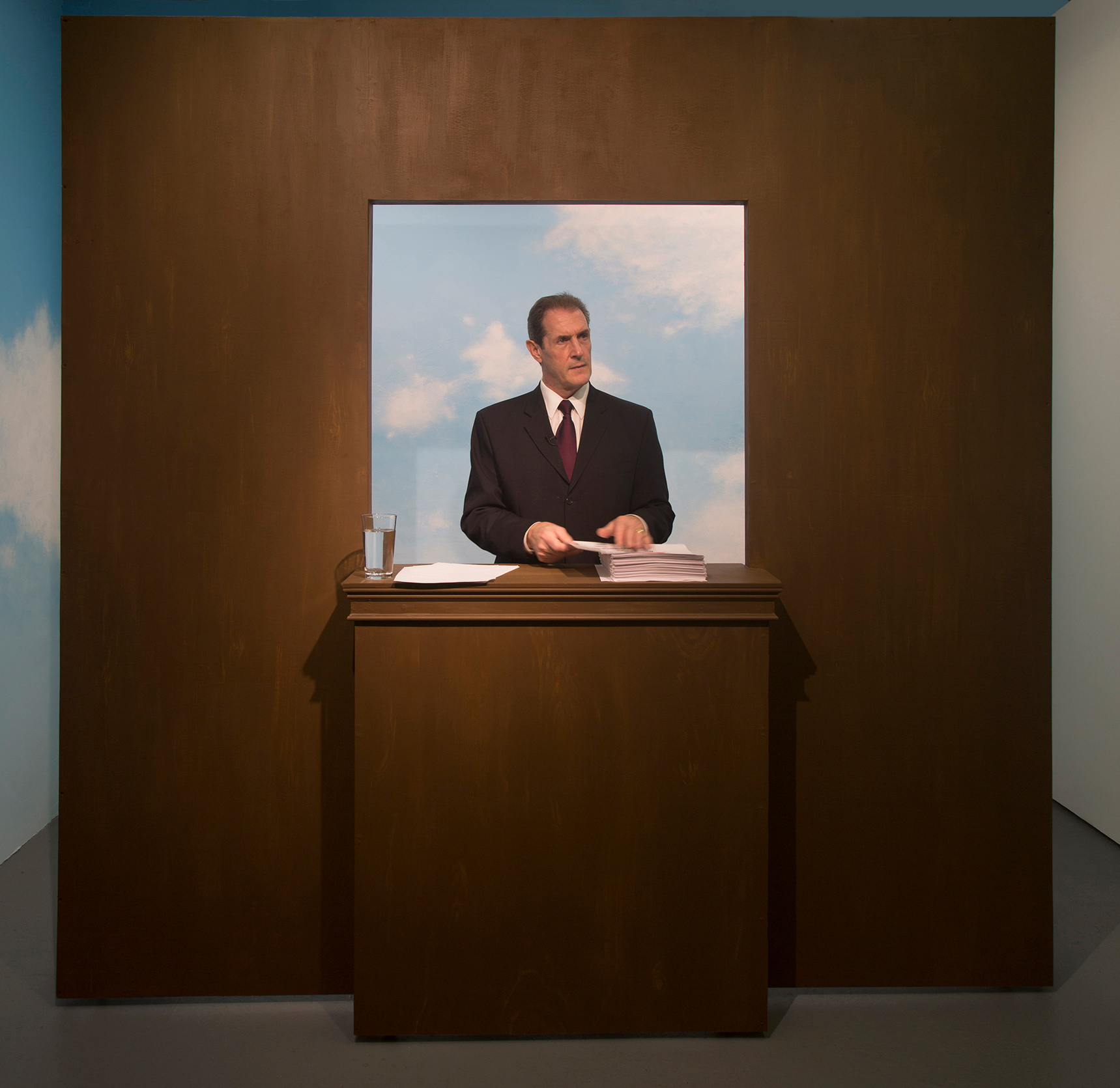 a zoomed out image of the entire scene that was taken during a performance of the project. The podium is in fact a large faux dark wood wall with a window cut out of its middle. Where the window is cut out a podium is attached (so that the wall and podium become one). In this image, we see the man standing behind the podium and window, looking to his left while grasping at a sheet of paper.