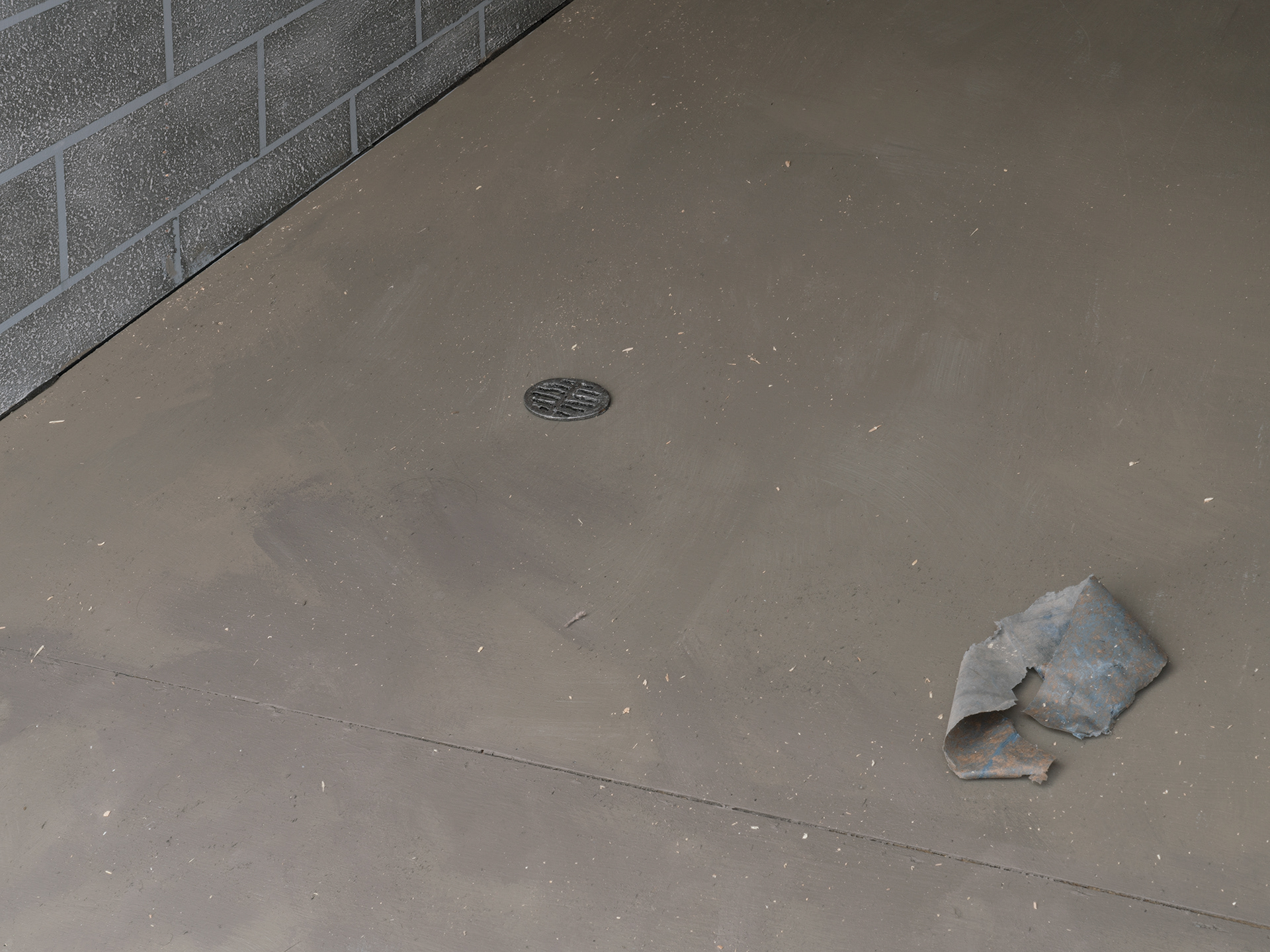 cropped in image of sculpture interior, showing a miniature drain on the grey floor to the middle left, and the piece of blue plastic trash to the right of the image