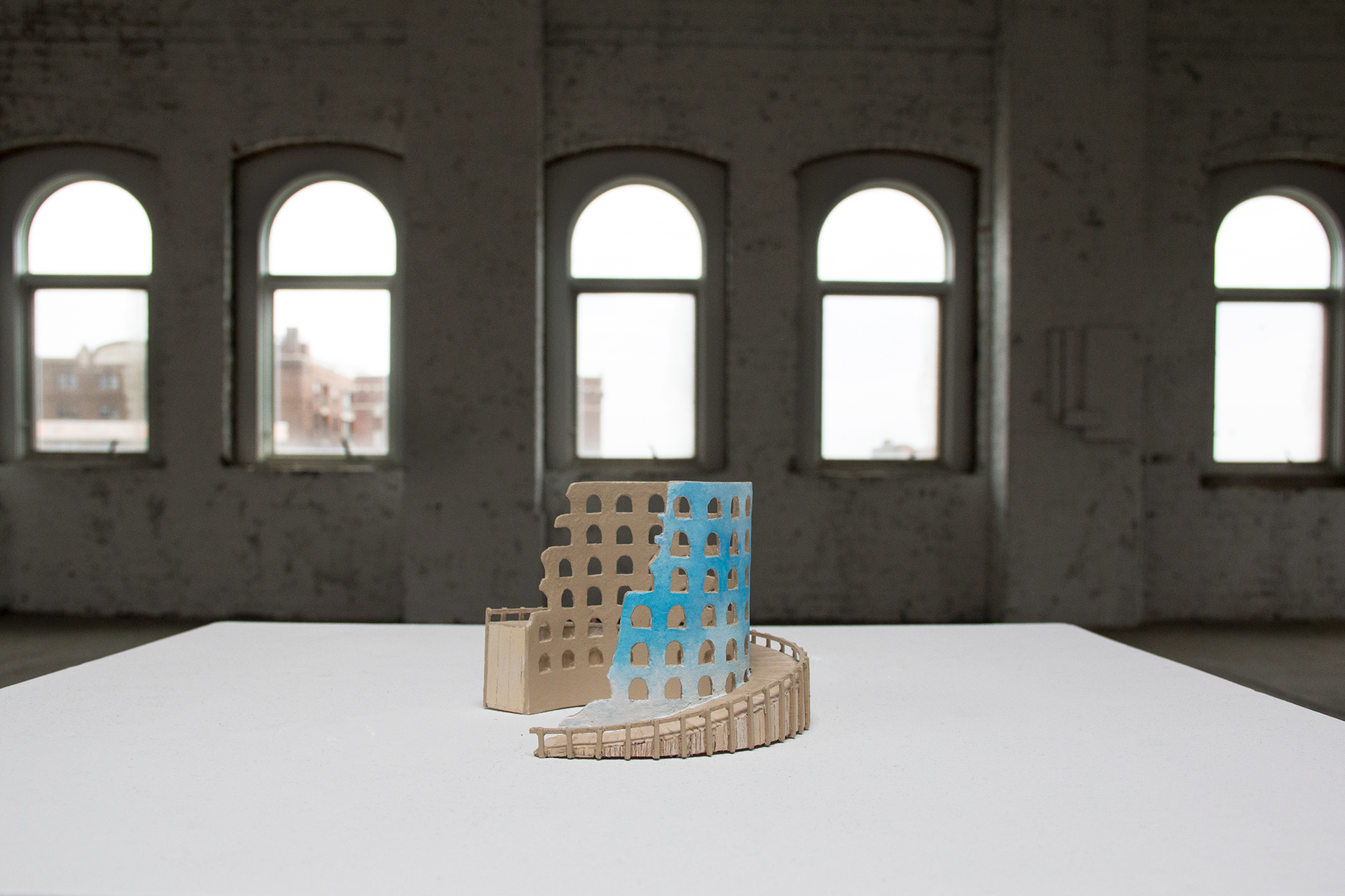 A small balsa wood model of a round Panopticon / Colosseum-like structure made as a ramp. The outer walls of the structure are painted with a blue sky and clouds. It sits on a white pedestal in a large industrial space.