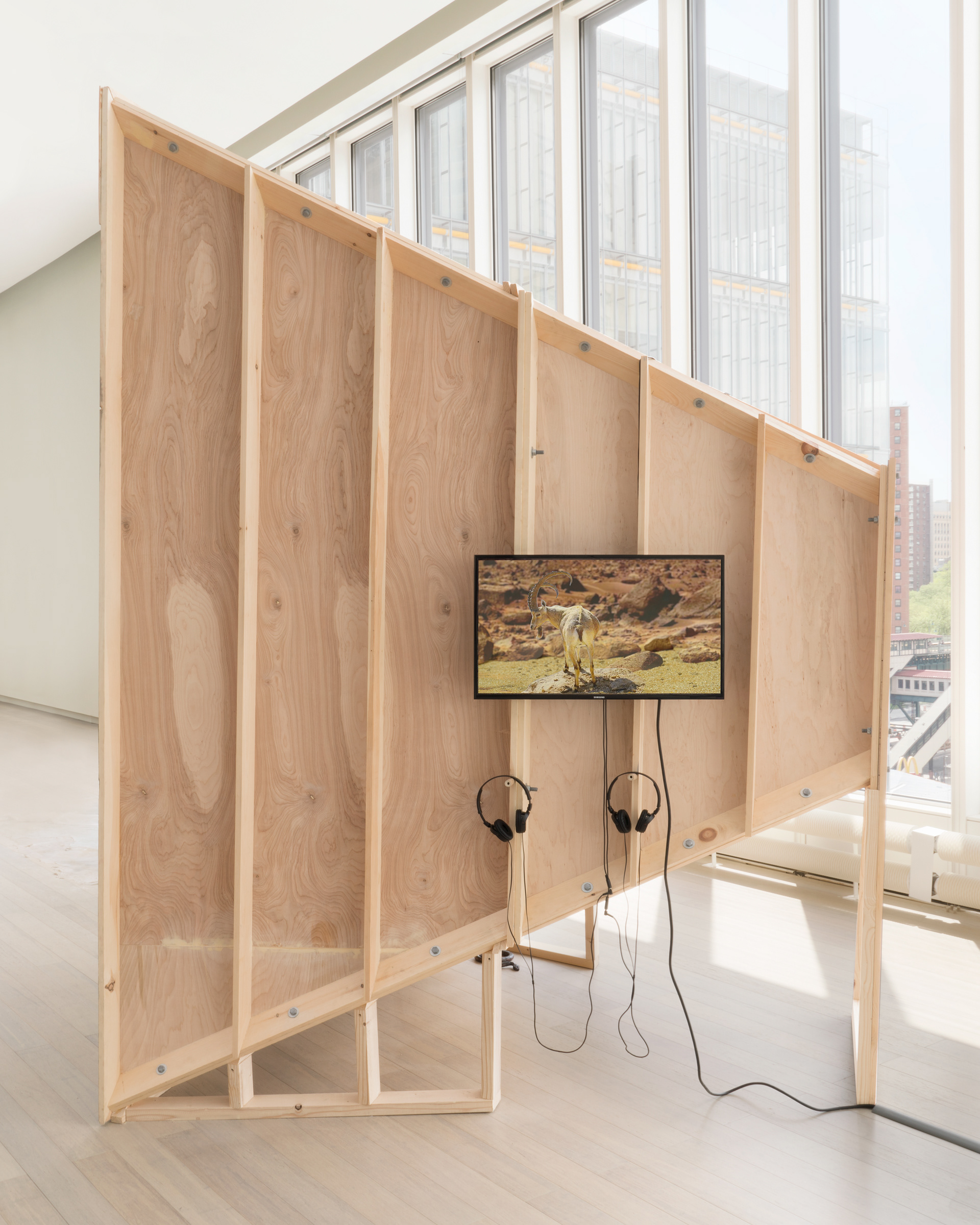 "Silver Bullet Test Site" detail from the right side of sculpture, showing the raw wood exterior of the structure, and a television mounted to the side with headphones. On the screen we see a faint image of an ibex standing in the desert.