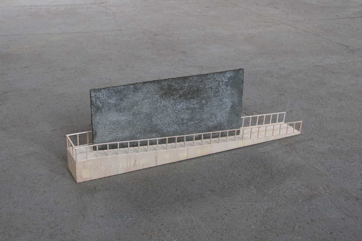 Balsa wood model using concrete separation wall with a ramp climbing up one side, around, and back down. The model sits on a concrete floor.