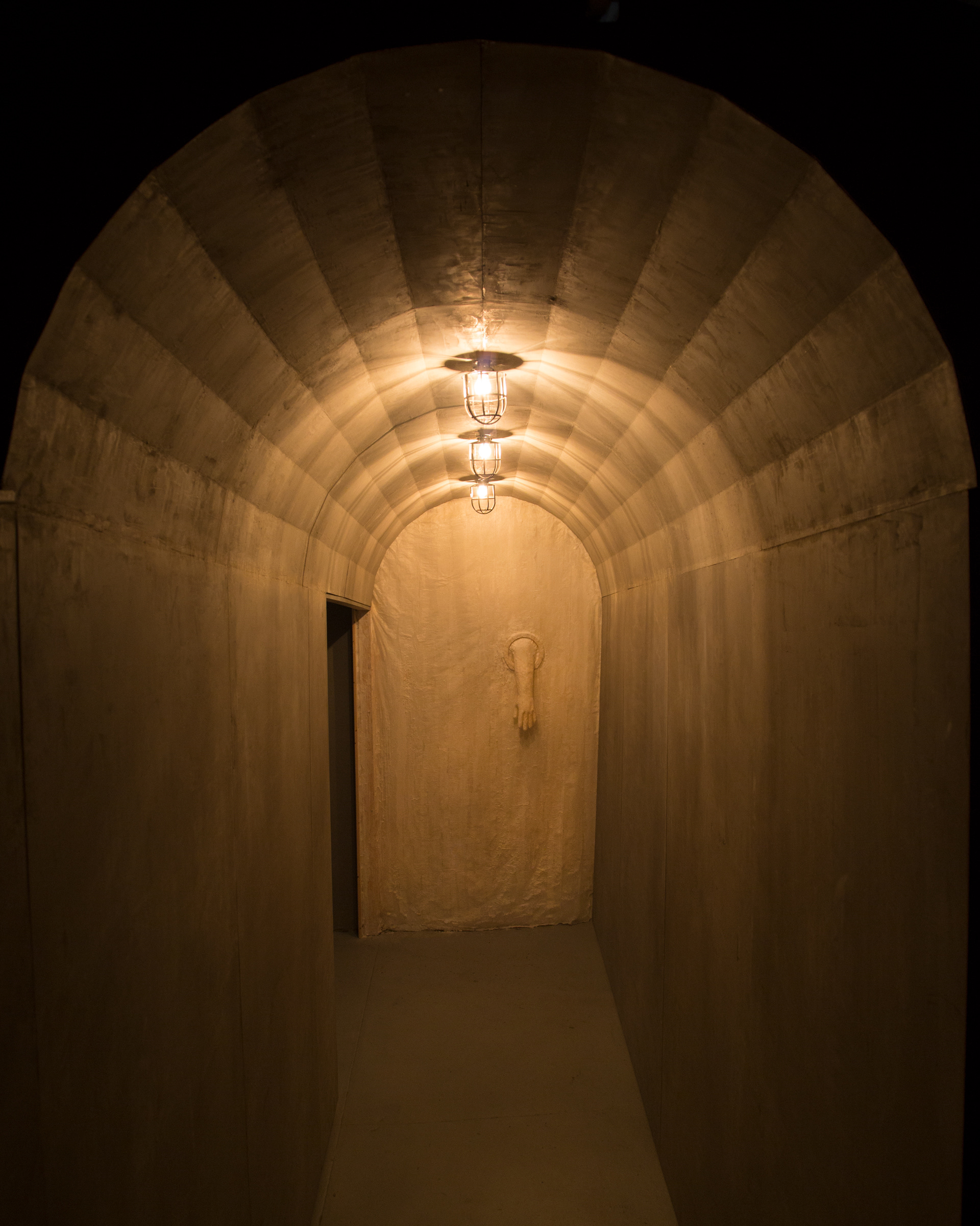 View from the stairway into the gallery. A tunnel-like structure at the gallery’s entry with cage lights above. A paper and beeswax membrane with a quarantine glove is at the end of the entry hallway. This image is lit with only lights from the installation (no outside or gallery light interruption)