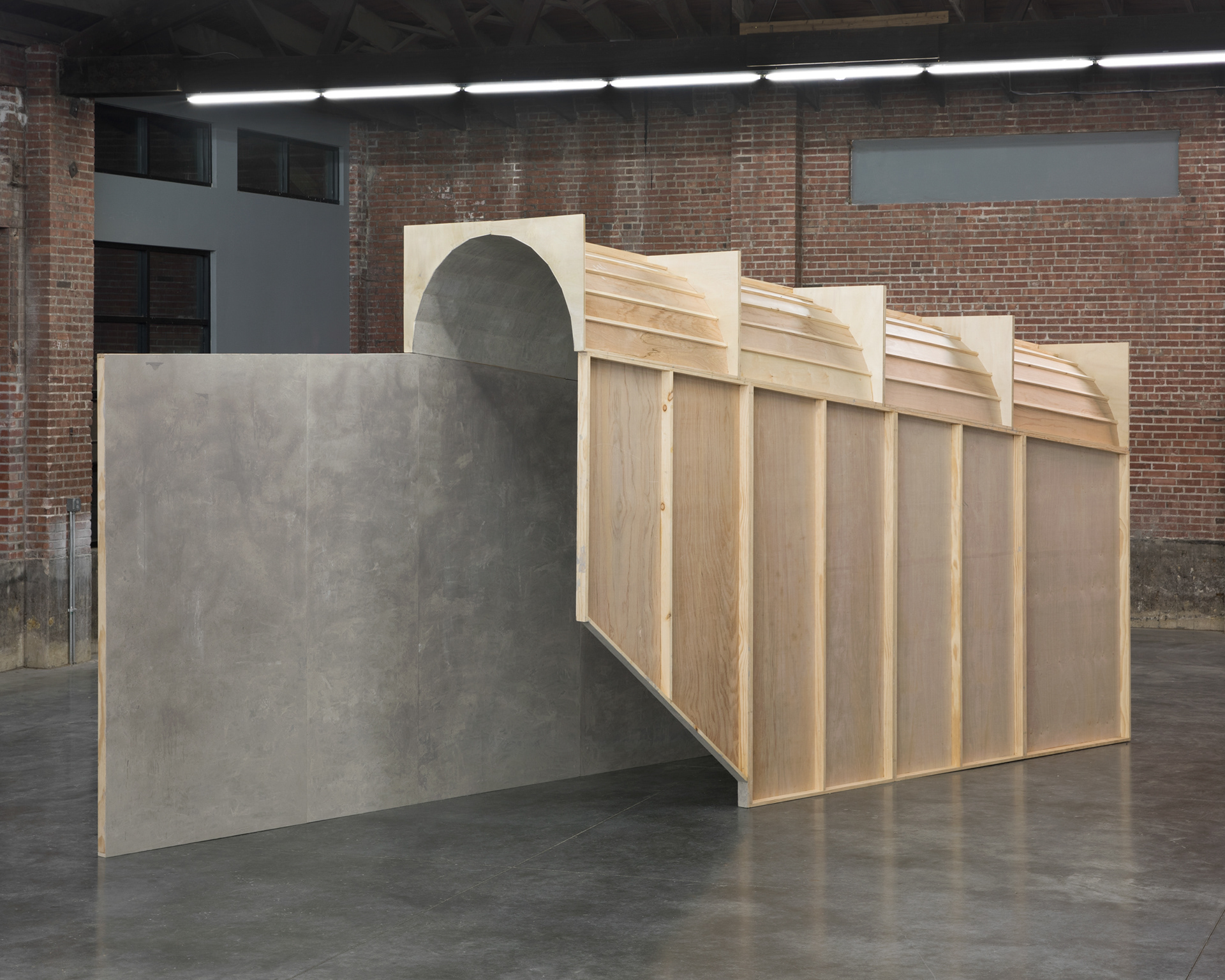 An angled view of a tunnel-like structure constructed of wooden panels standing in a large industrial space. The space has concrete floors, red brick walls, and an arched ceiling.