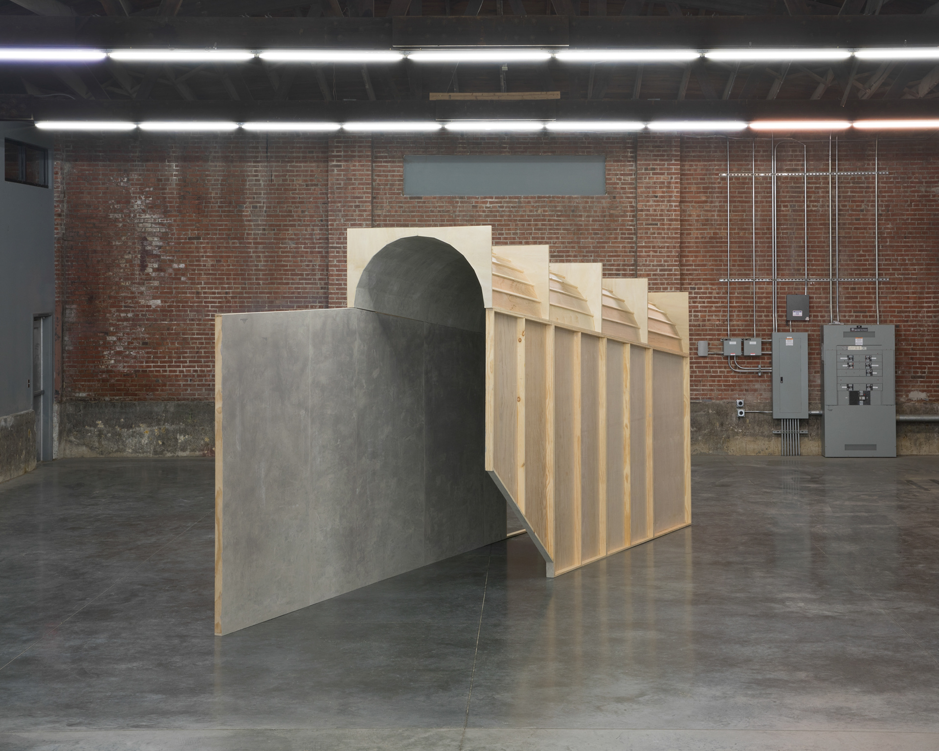 Frontal view of a tunnel-like structure constructed of wooden panels standing in a large industrial space. The space has concrete floors, red brick walls, and an arched ceiling.