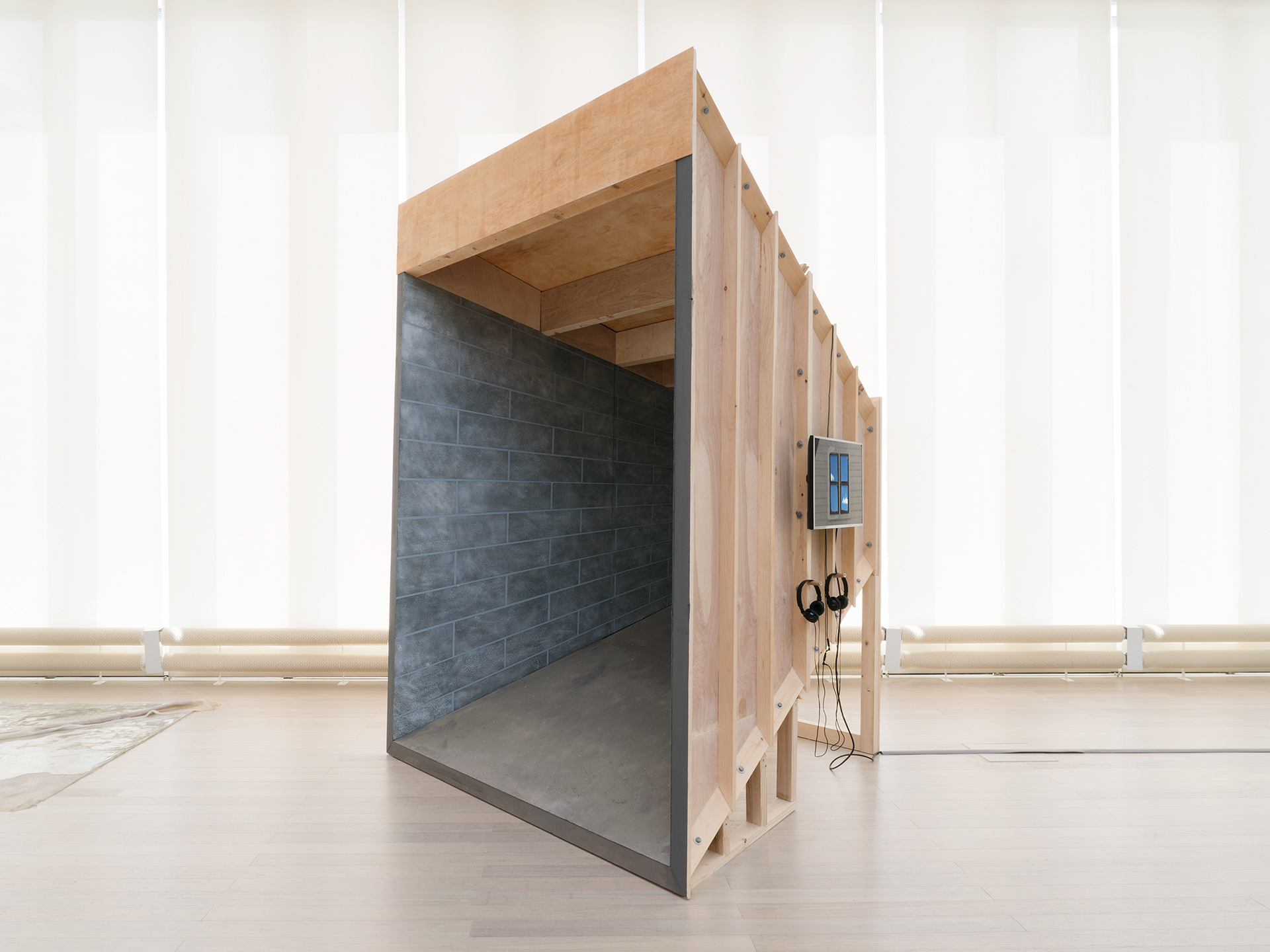 "Silver Bullet Test Site" overall shot. image description: A sculpture stands in front of a large row of windows (shades closed). The sculpture is a point perspective room made out of wood, and is painted to resemble a basement on the interior. a monitor hangs on the outside of the structure.