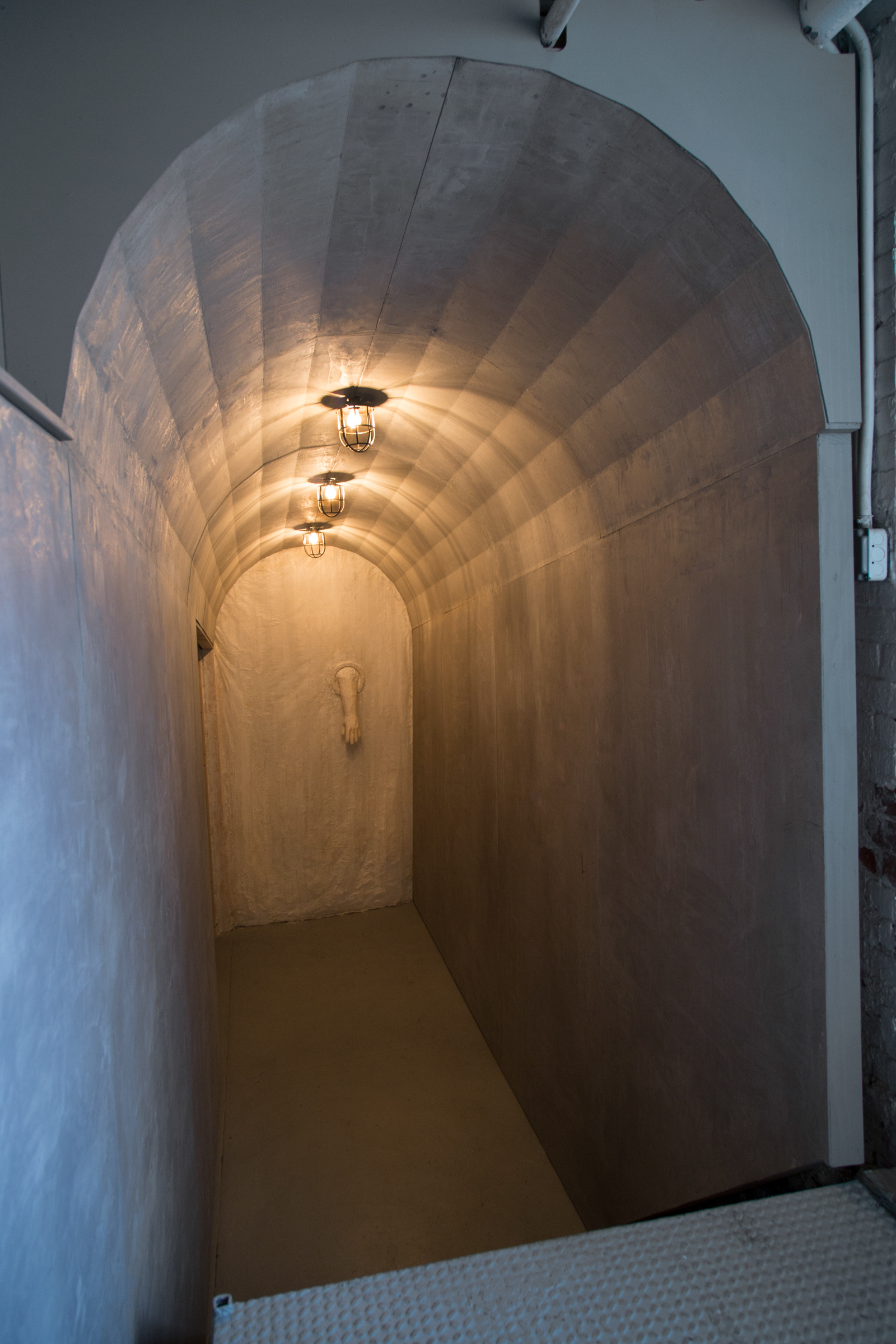 angled view of a tunnel-like structure at the gallery’s entry with cage lights above. A paper and beeswax membrane with a quarantine glove is at the end of the entry hallway.