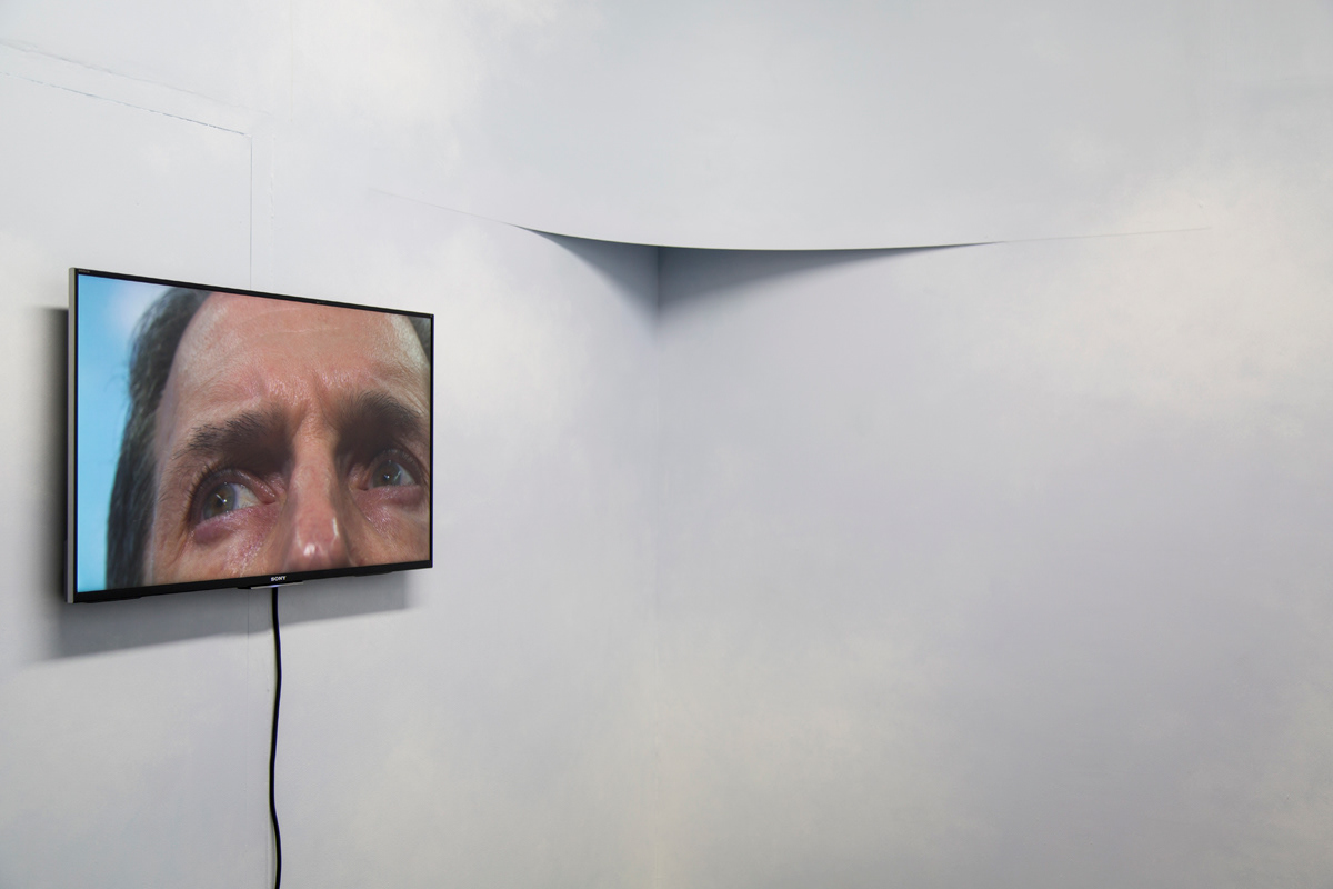 A detail shot of the TV monitor with the middle ages man's eyes in the frame. To the right of the TV you can see the border of the room with a rounded edge.