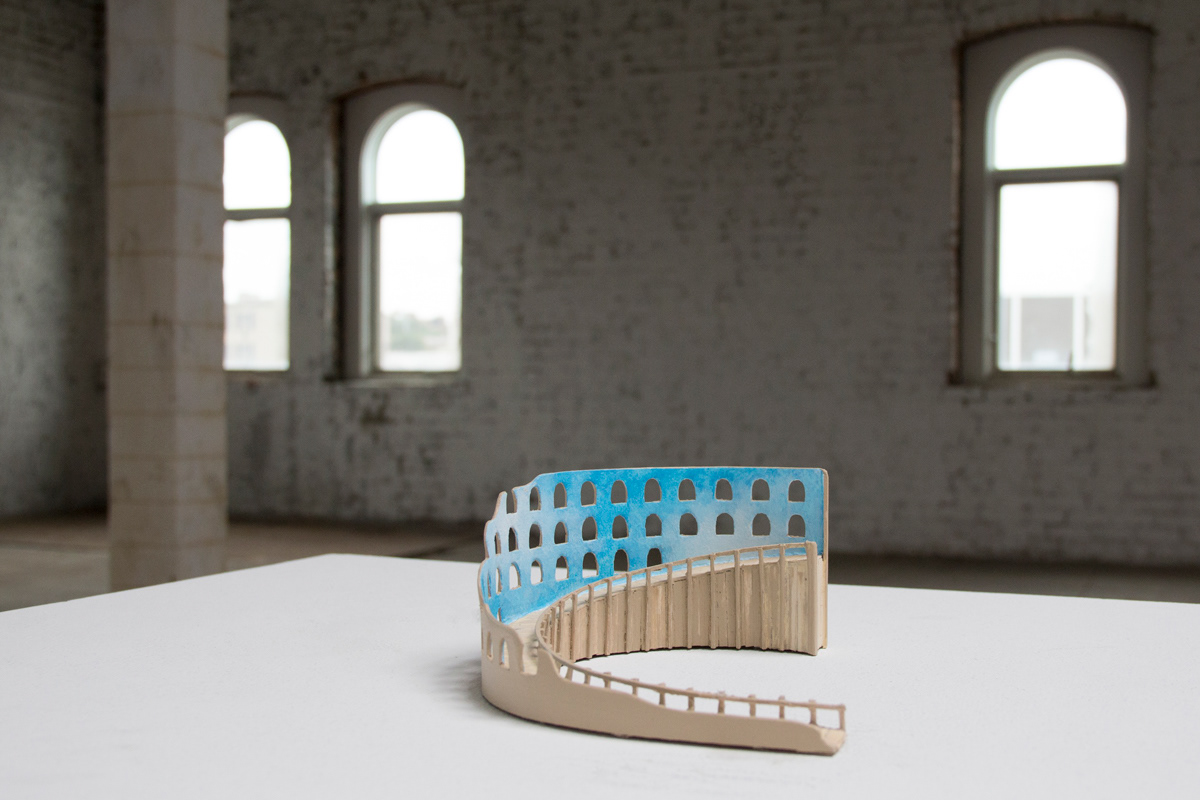 A small balsa wood model of a round Panopticon / Colosseum-like structure made as a ramp. The inner walls of the structure are painted with a blue sky and clouds. It sits on a white pedestal in a large industrial space.