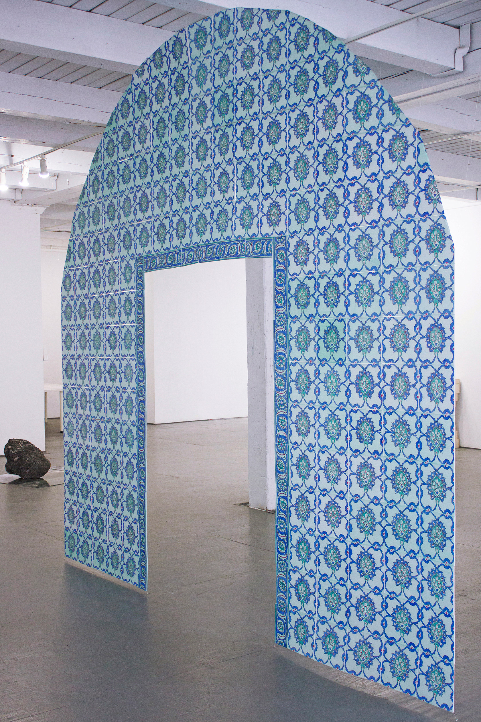 Angled view of "Their Natural State," a sculpture recreation of an archway facade at King Davids Tomb in Jerusalem. The sculpture is made up of paper tiles created with vellum, hand drawn with marker, and strung together with fishing line. the final product is a ghost-like memorial. Each tile is painted with blue, turquoise, green and red floral patterns.