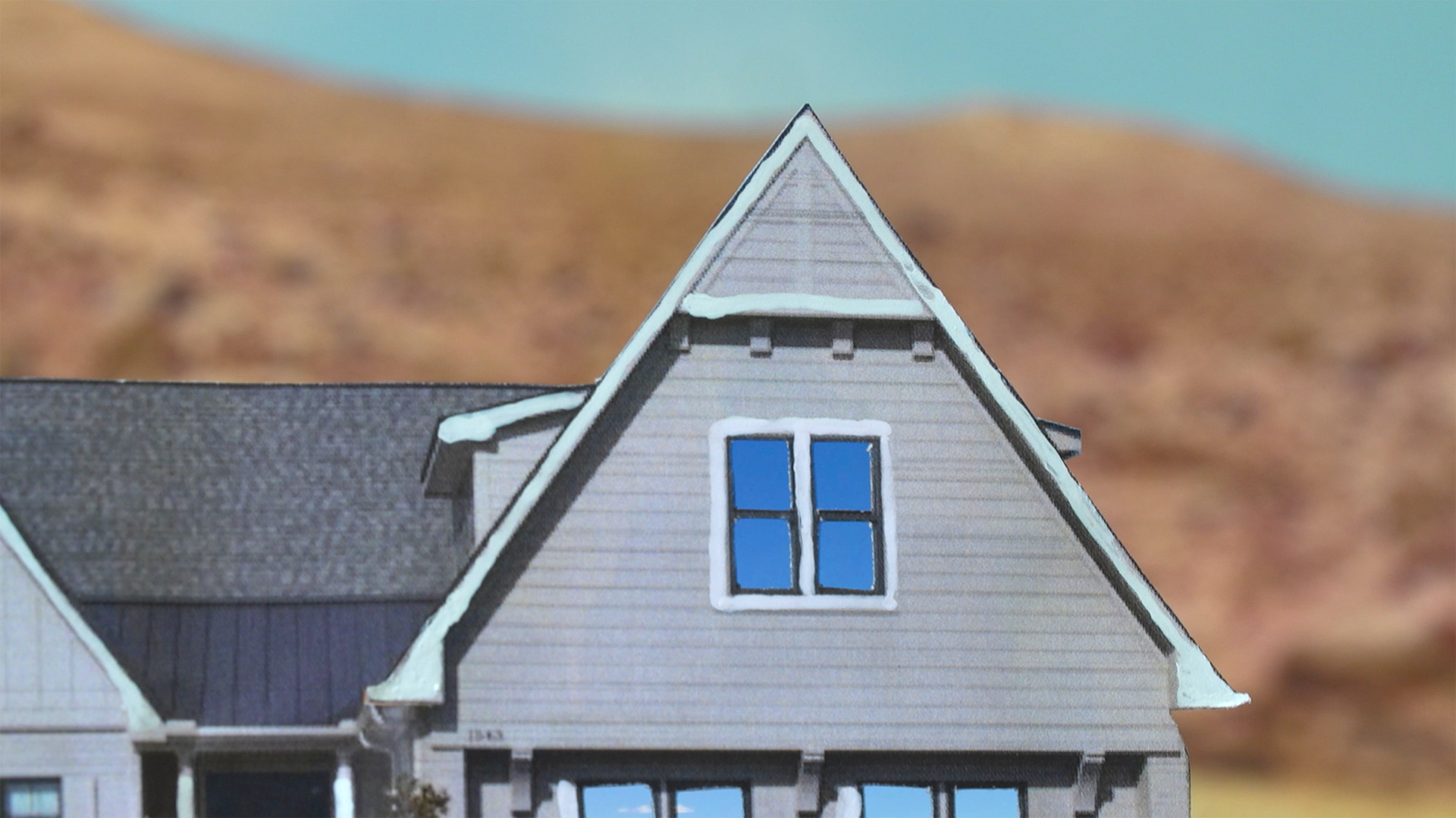 A video still taken from the "Silver Bullet Test Site" video. Image description: a close-up view of a paper house with desert background. The windows reflect the sky. The image is created using found online images that are cut and assembled for the video.