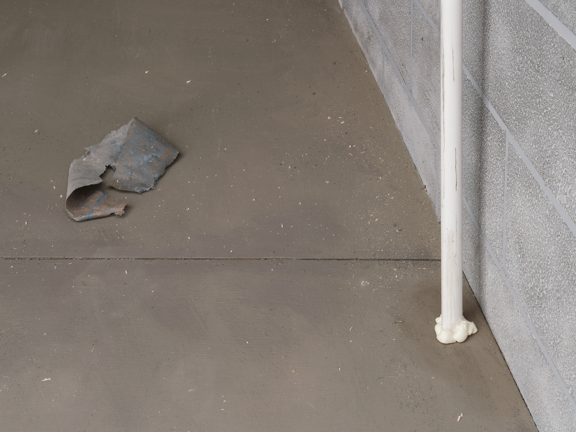 a close up of the sculpture's interior showing a piece of blue plastic trash on the grey floor to the left of the image, and a detail of a white plastic pipe coming out of the floor with spray foam at its base on the right.