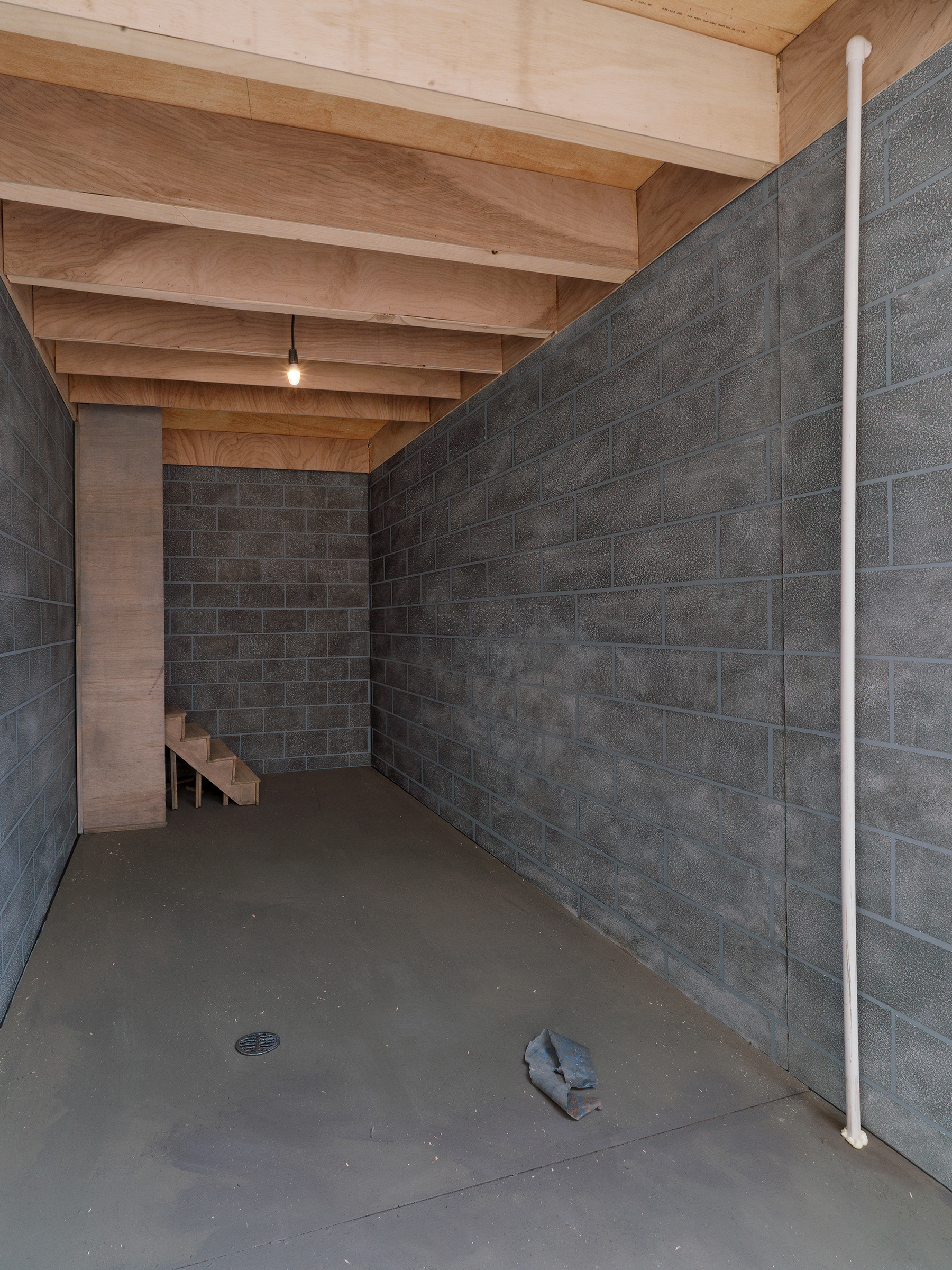 detail image of the interior of "Silver Bullet Test Site", including painted faux cinderblock walls, a miniature staircase to the back, a white pipe moving from floor to ceiling is to the right of the image. There is also what appears to be some trash on the floor at the bottom mid-right of the image, and a small drain to the left.