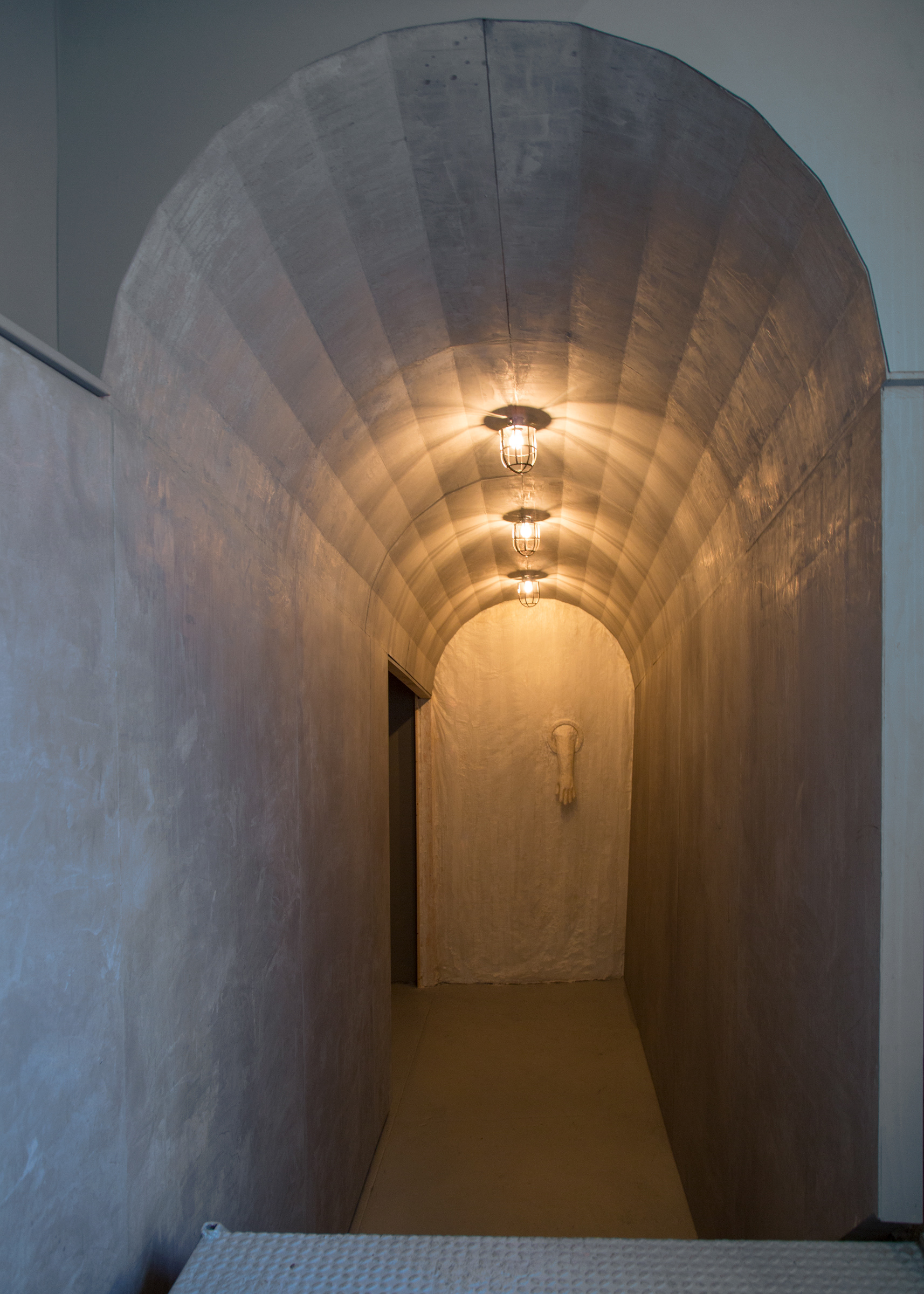 Frontal view of a tunnel-like structure at the gallery’s entry with cage lights above. A paper and beeswax membrane with a quarantine glove is at the end of the entry hallway.