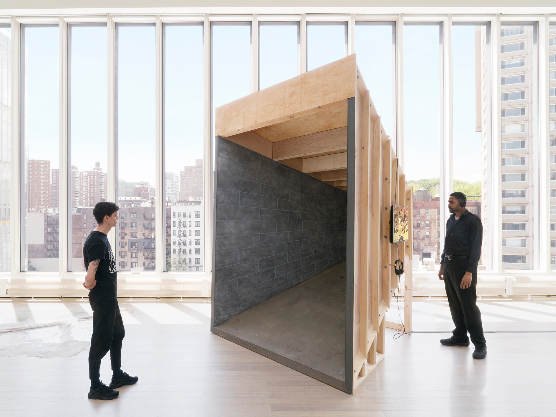 "Silver Bullet Test Site" overall shot (shades open) with two viewers interacting with the sculpture: one stands in front contemplating the entry to the room, and the other to the side of the sculpture, using headphones and watching the video on the exterior of the structure.
