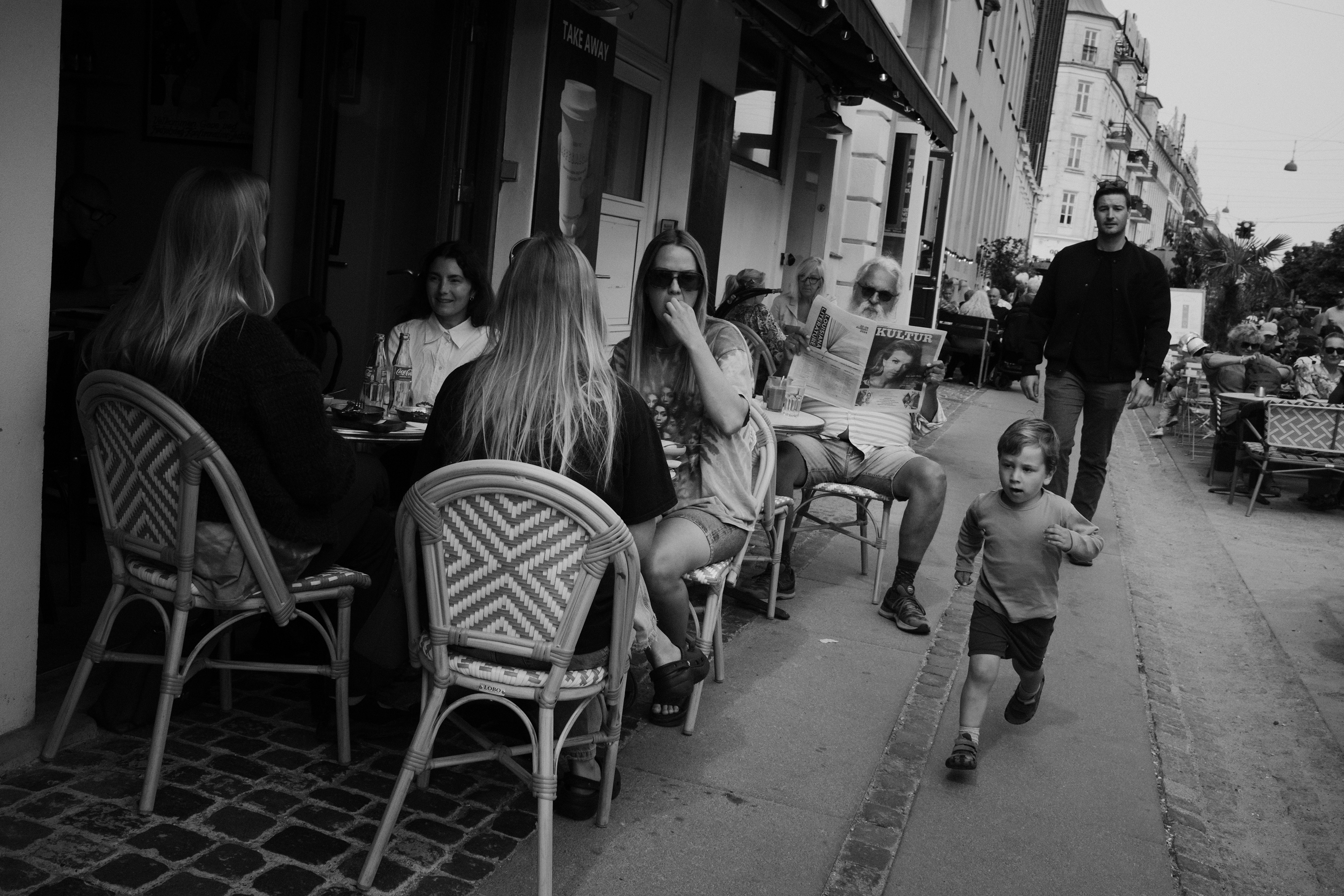 Child runs past people at a café. Copenhagen, Denmark 2024.
