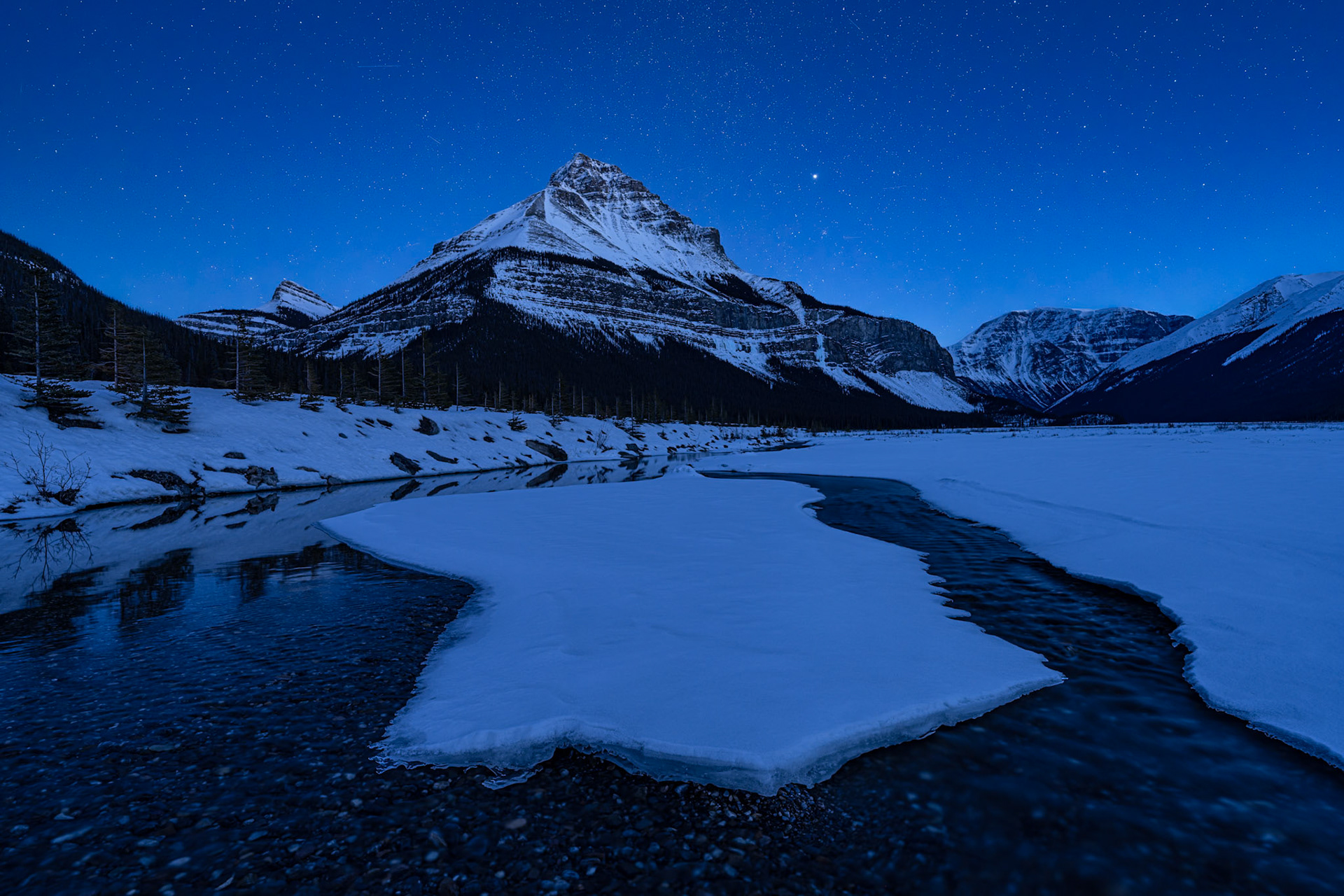 A large piece of ice floats along a partially frozen creek beneath Tangle Peak and a star-filled sky in the Canadian Rockies.