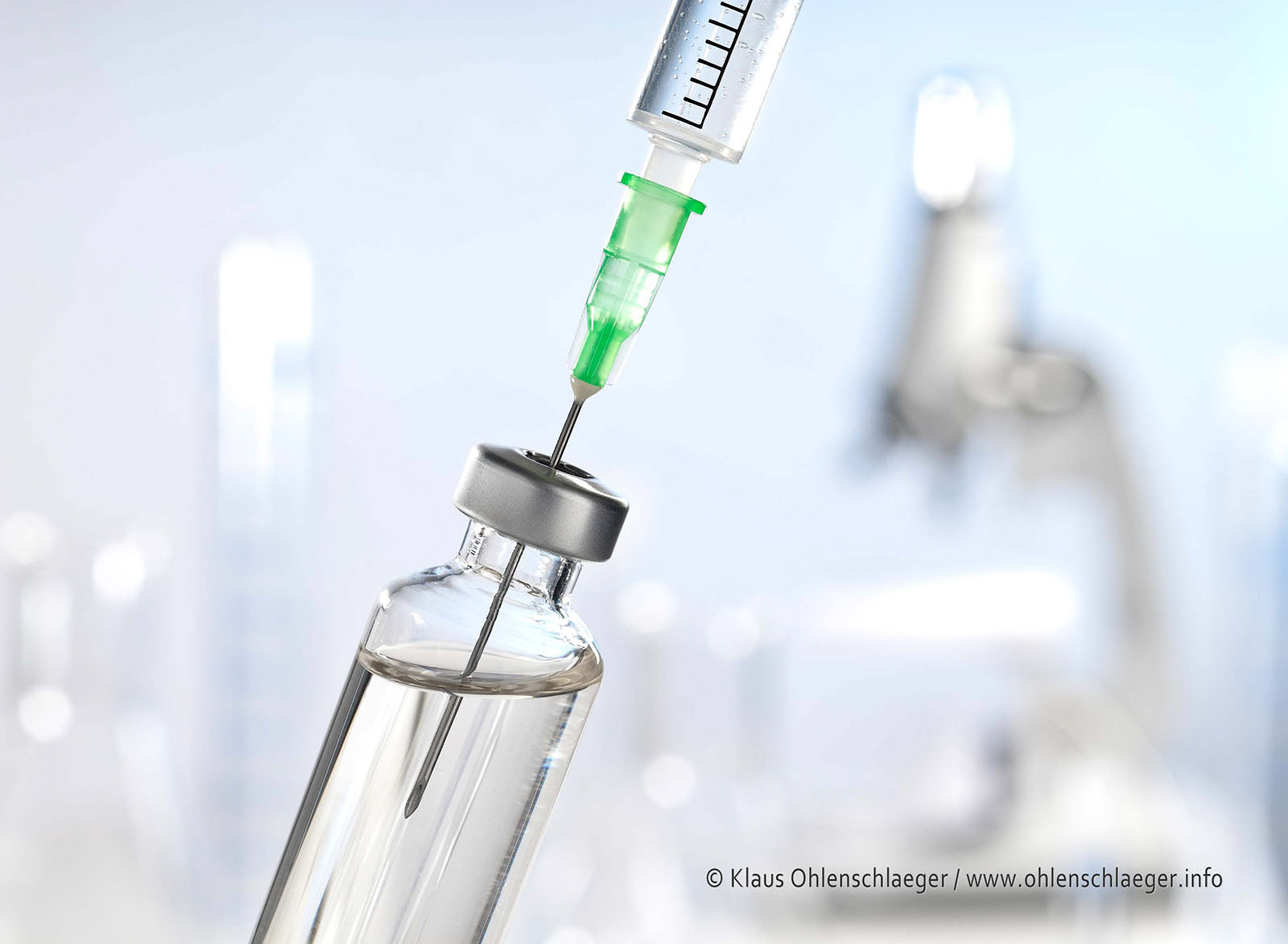 Laborszene mit Spritze und Medizin im Fläschchen im Vordergrund und Mikroskop und weiteren Utensilien im Hintergrund