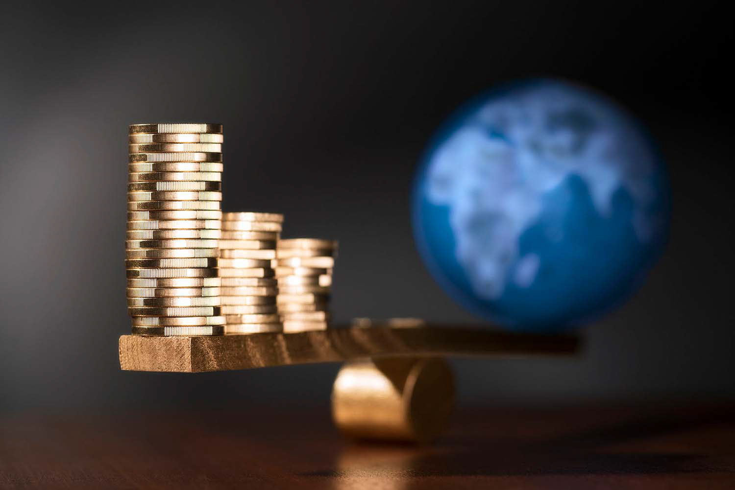 Konzept Geld regiert die Welt. Stapel aus Münzen im Vordergrund und Weltkugel hinten auf einer goldfarbenen Wippe. Studio für Produktfotografie in Kronberg im Taunus - Fotograf Klaus Ohlenschläger - Frankfurt - Kronberg - Bad Homburg